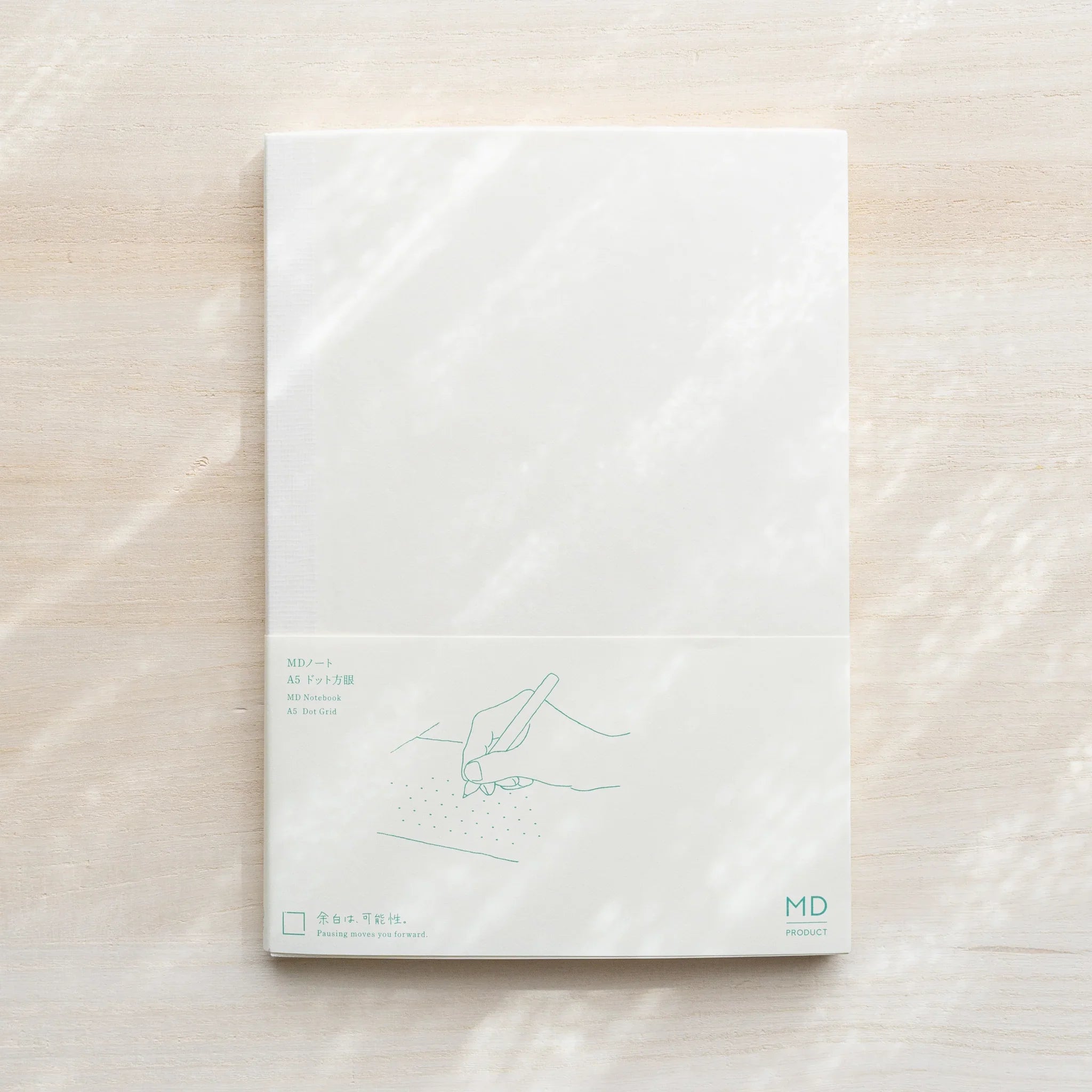 Midori MD Notebook A5 Dot Grid in glassine wrapping with belly band showing hand illustration and MD logo on light wood
