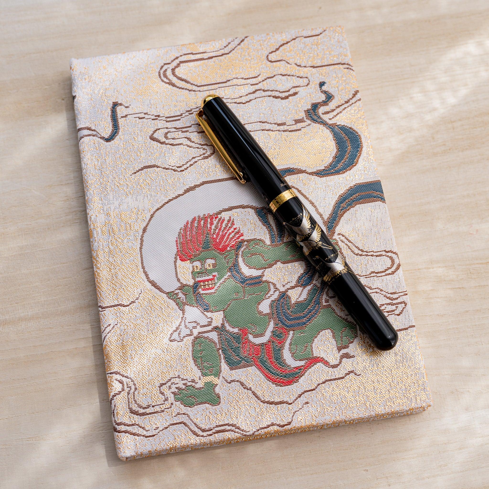 Notebook front cover with green wind god Fujin in woven textile, black fountain pen resting on top