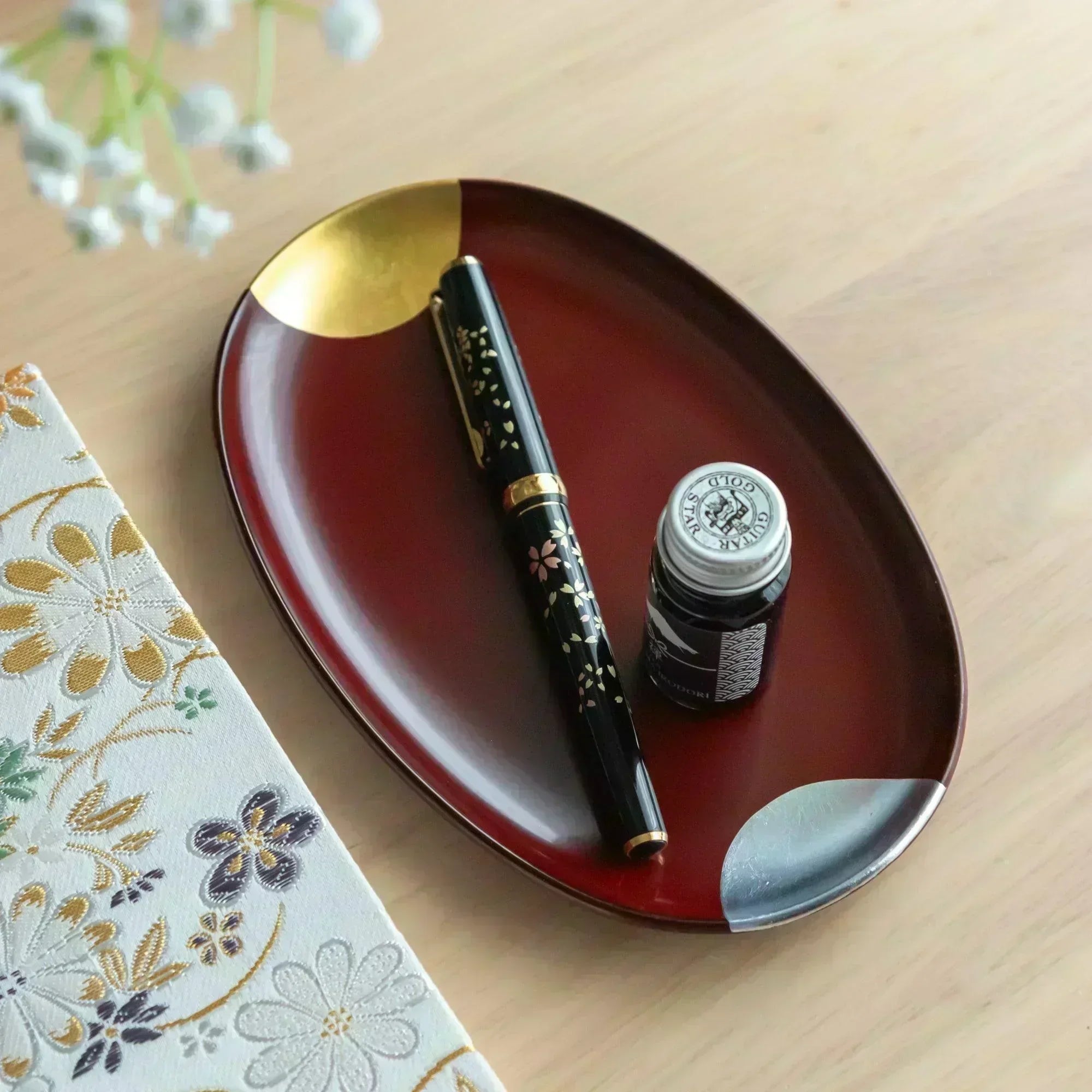 A Japanese stationery setup featuring a black sakura pen and ink bottle on a red lacquer tray with gold and silver accents.