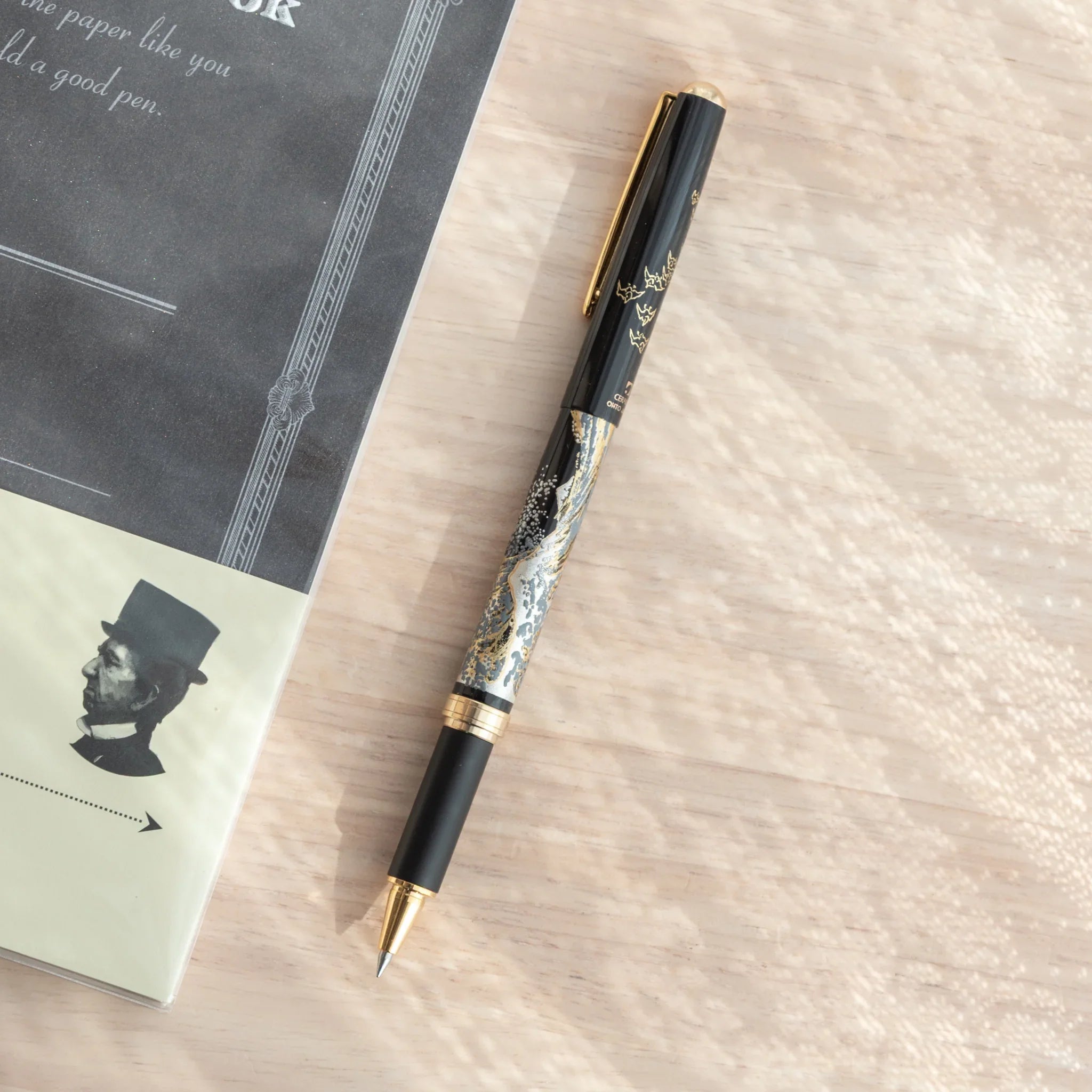 Maeju Yamanaka ballpoint pen with Great Wave silkscreen in silver and gold on black barrel, beside dark gray notebook on wood
