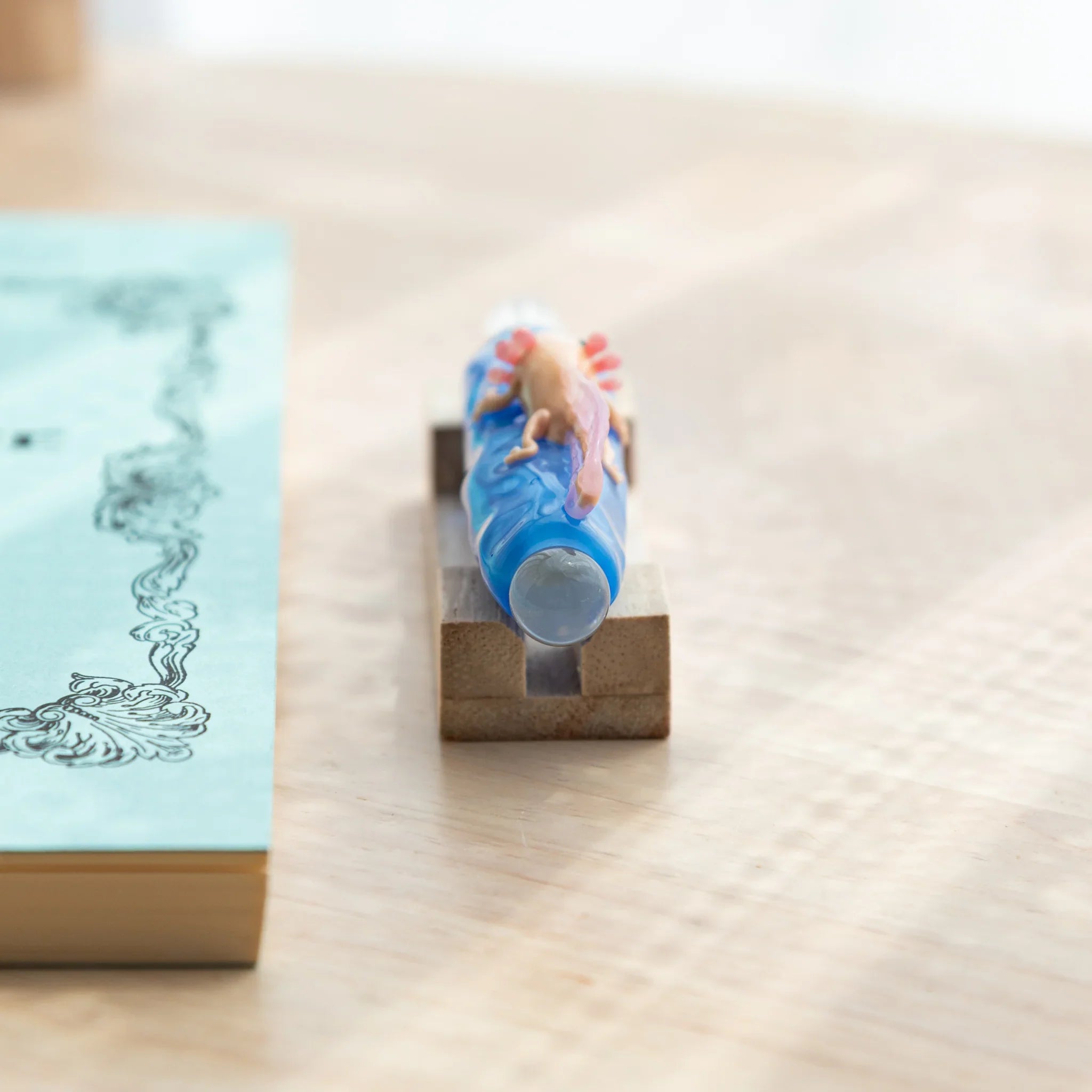 Blue glass dip pen with a small pink-gilled axolotl on the shaft, placed on a wooden stand beside a light blue ornate notebook.