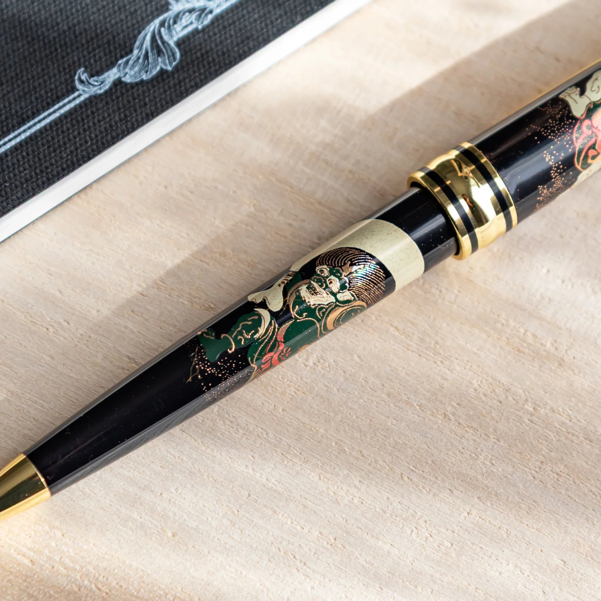 Japanese Ballpoint Pen on beige cloth with gold & black Raijin and Fujin design, beside a dark notebook.