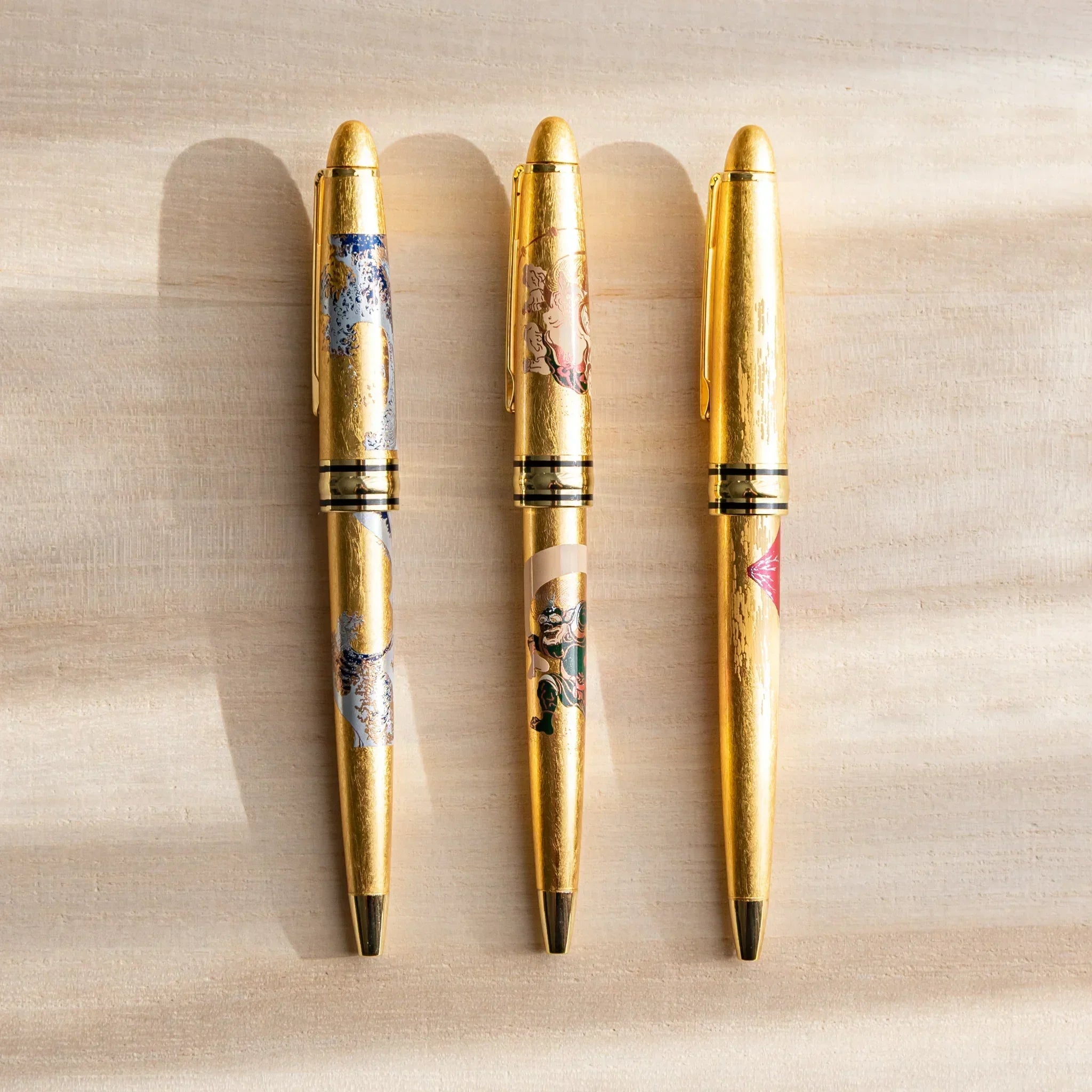 Three gold leaf Japanese Ballpoint Pens with Raijin and Fujin artwork displayed on a light wood surface.