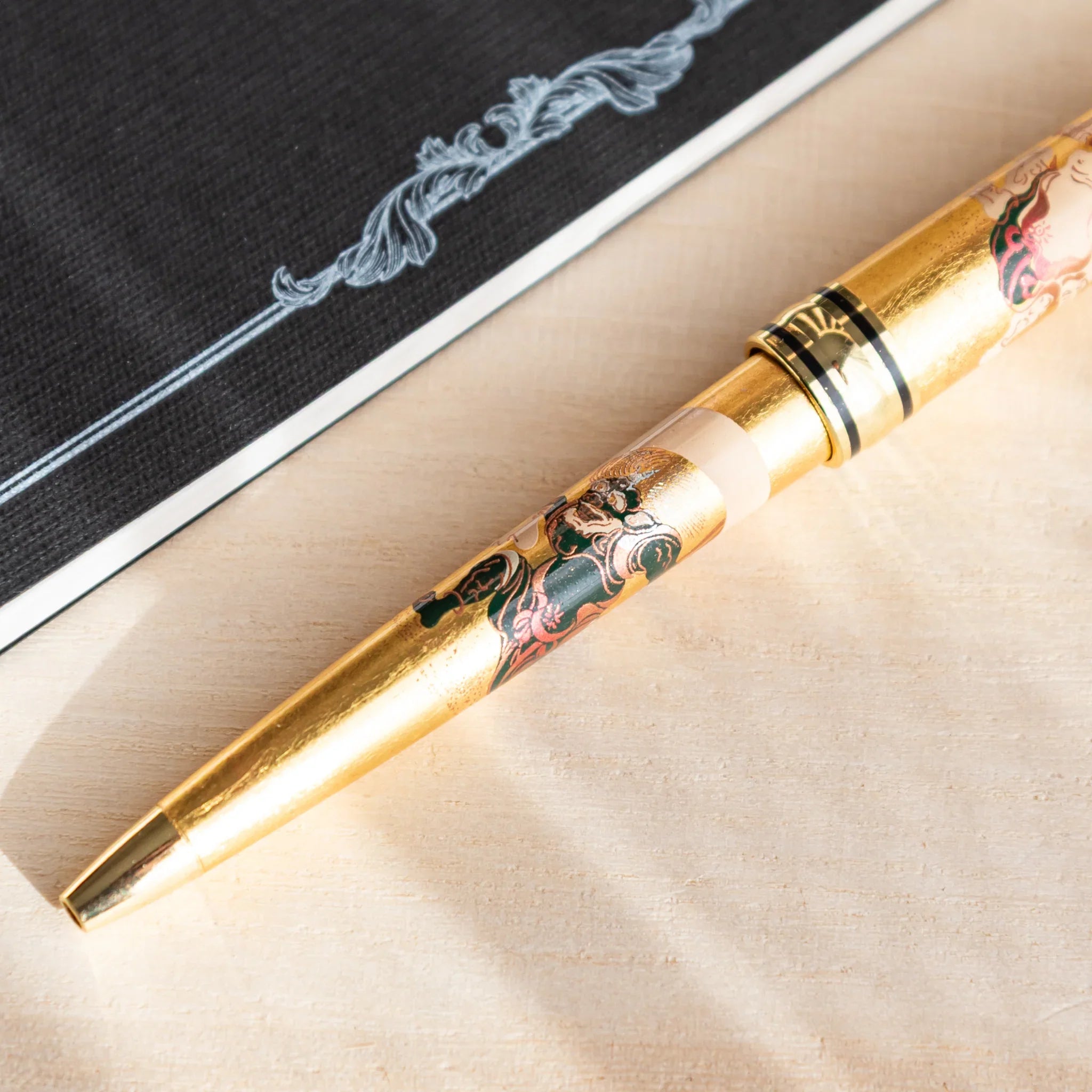 Japanese Ballpoint Pen with gold leaf Fujin design resting diagonally on a light wood desk beside a black notebook.