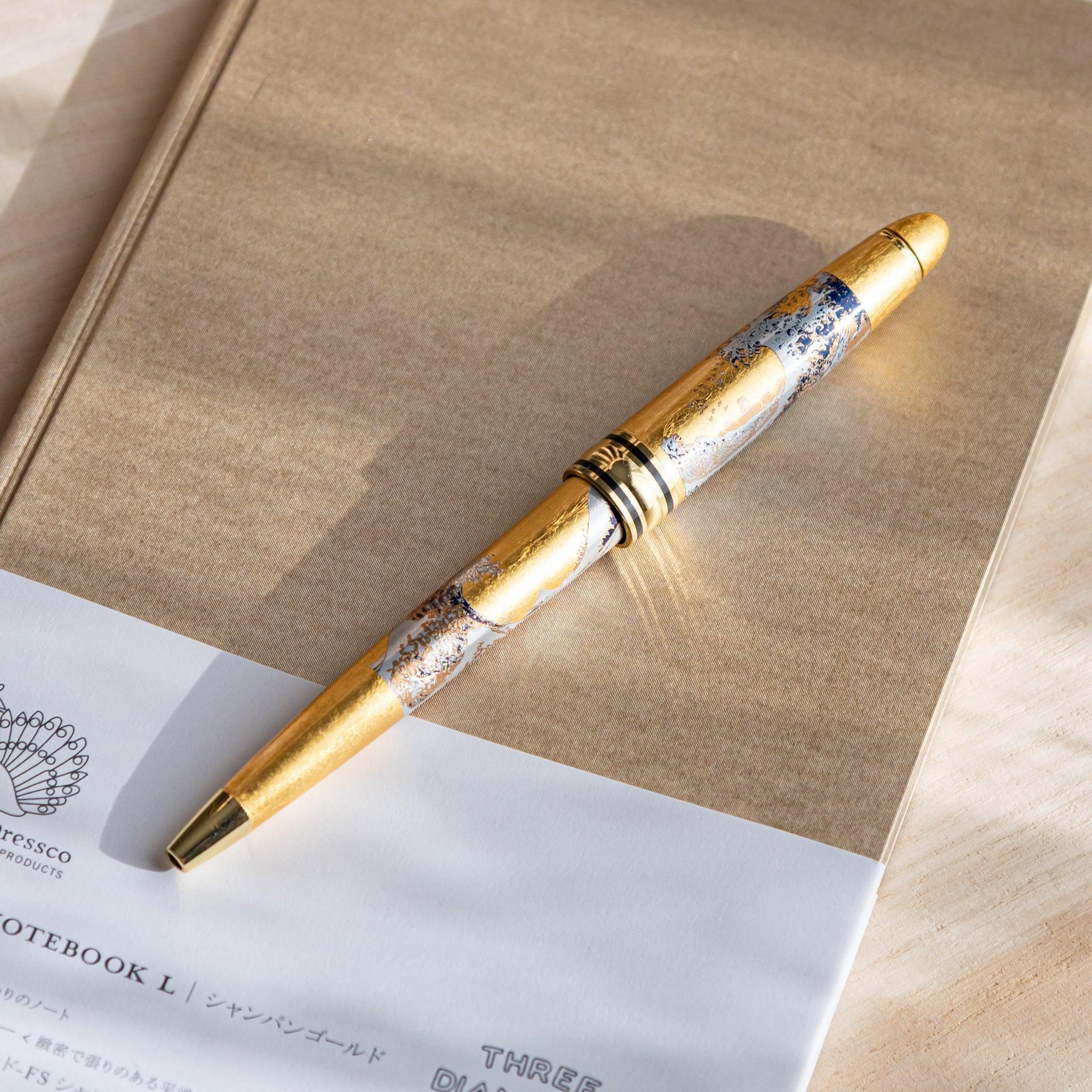Gold Leaf Great Wave Japanese Ballpoint Pen placed diagonally on a beige notebook, showing gold body with wave artwork.