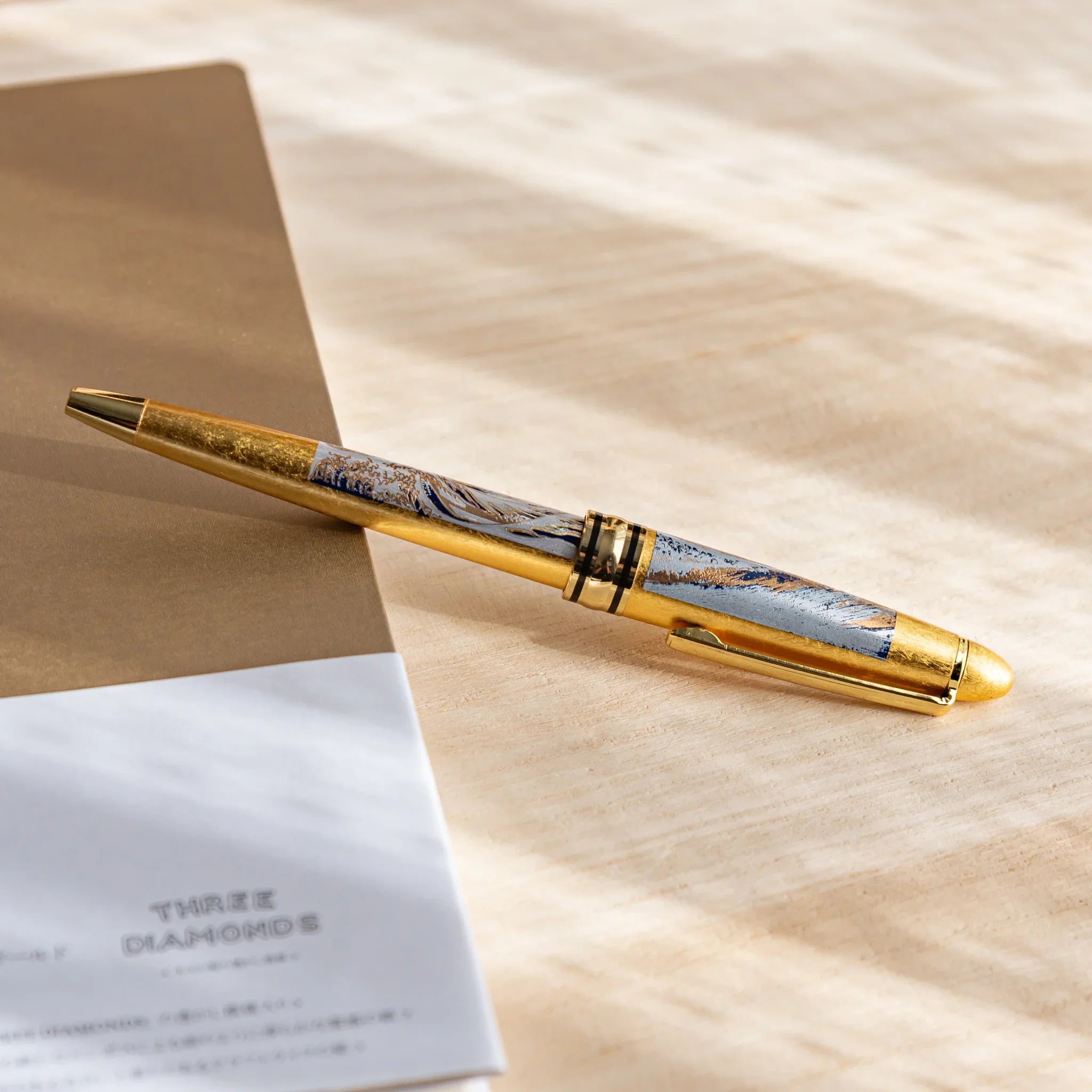 Gold Leaf Great Wave Japanese Ballpoint Pen on a beige and white notebook stack, showing blue-and-gold wave details and a gold body.