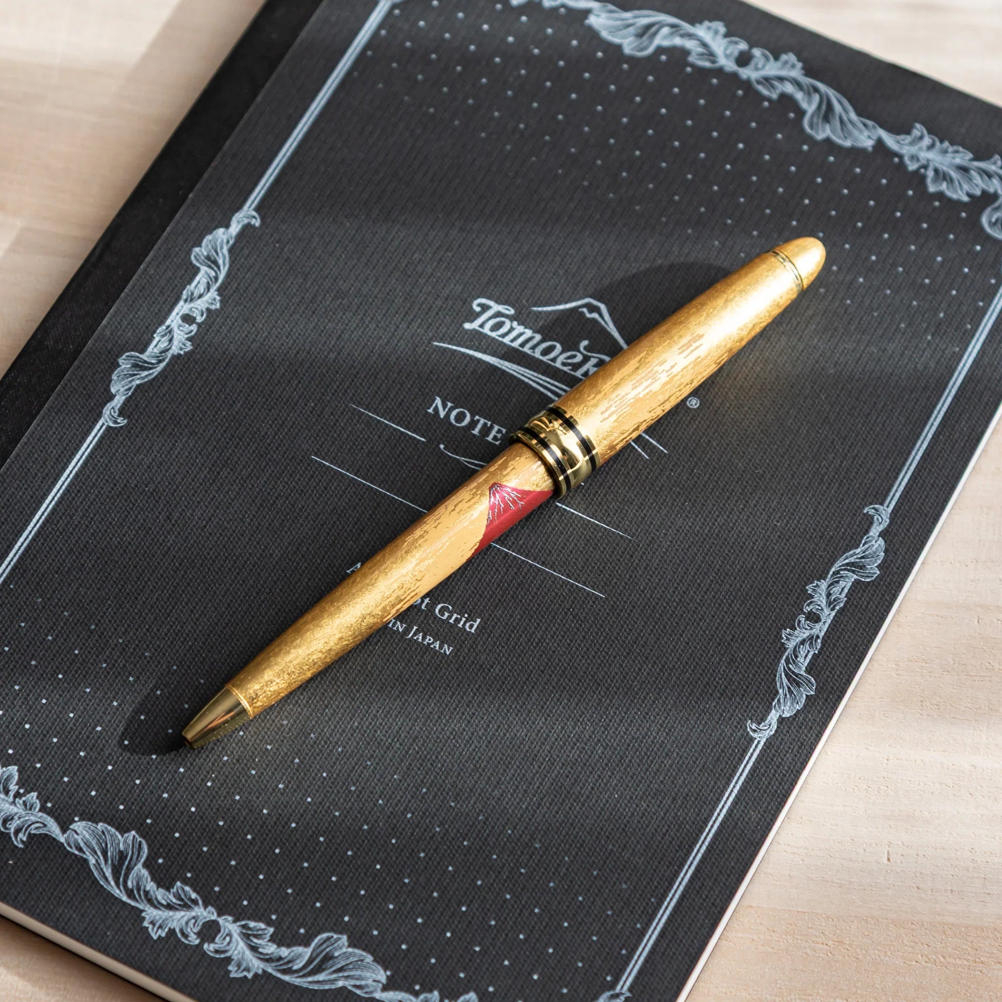 Gold Leaf Mt. Fuji Japanese Ballpoint Pen resting on a black Tomoe River notebook, showing red Fuji artwork and a gold body.