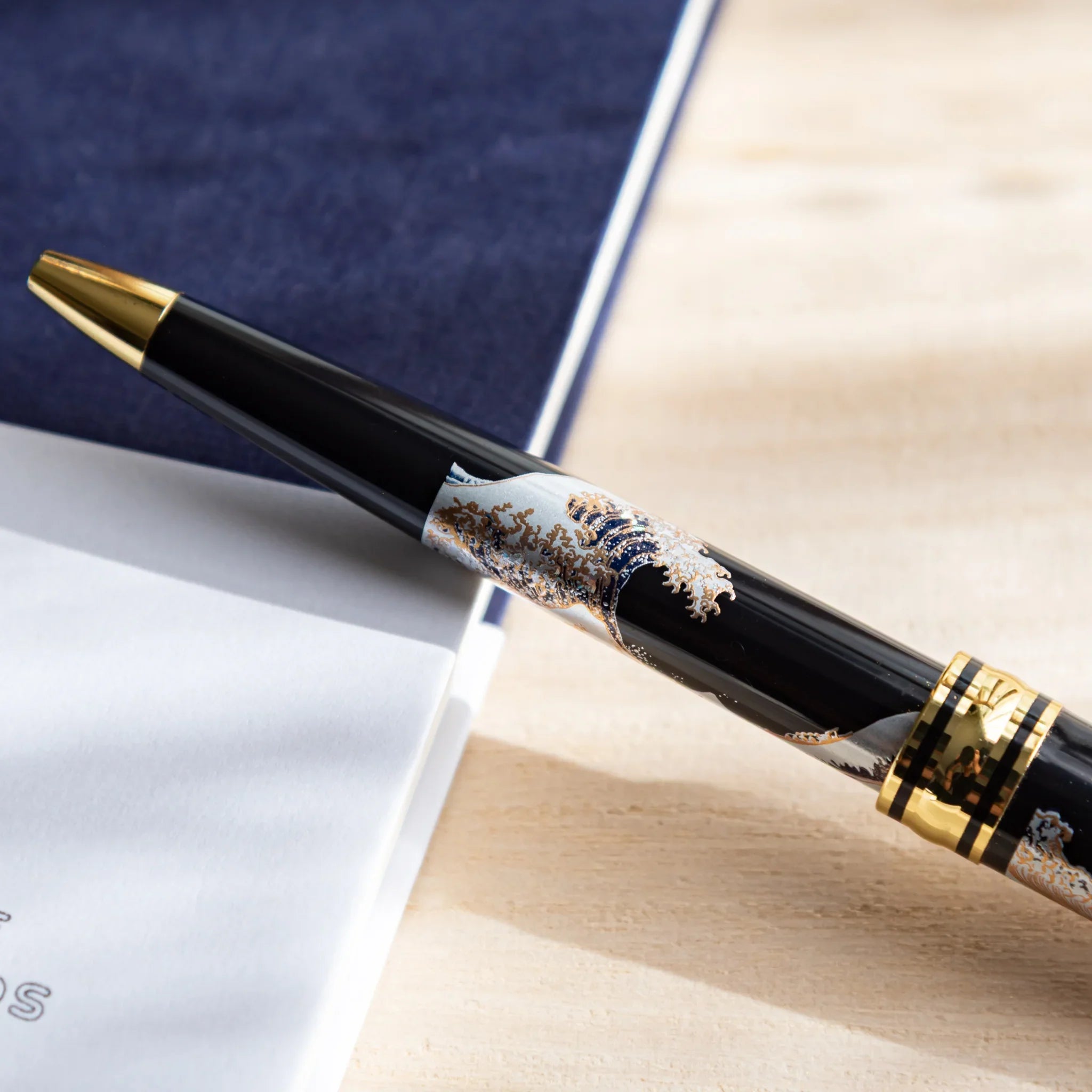 Japanese Ballpoint Pen with Great Wave design placed diagonally on the desk and notebook, showing black lacquer body.