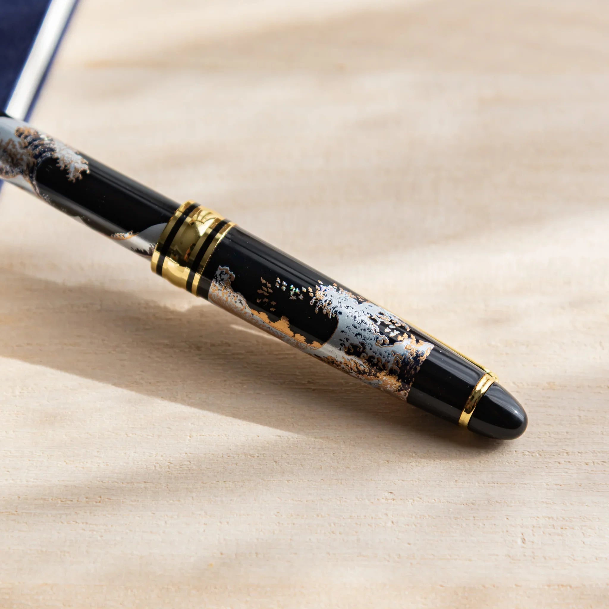 Japanese Ballpoint Pen with Great Wave design on a wood desk, showing black lacquer body, blue wave artwork.