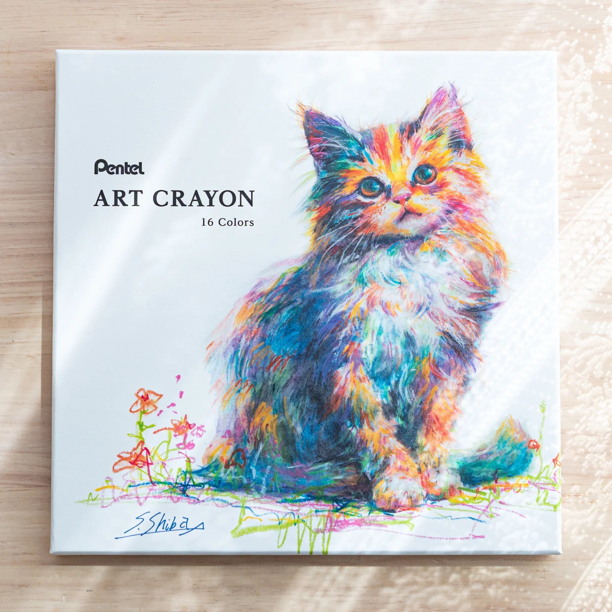 White square Pentel ART CRAYON 16 Colors box front with colorful fluffy kitten artwork signed S. Shiba on a wood surface
