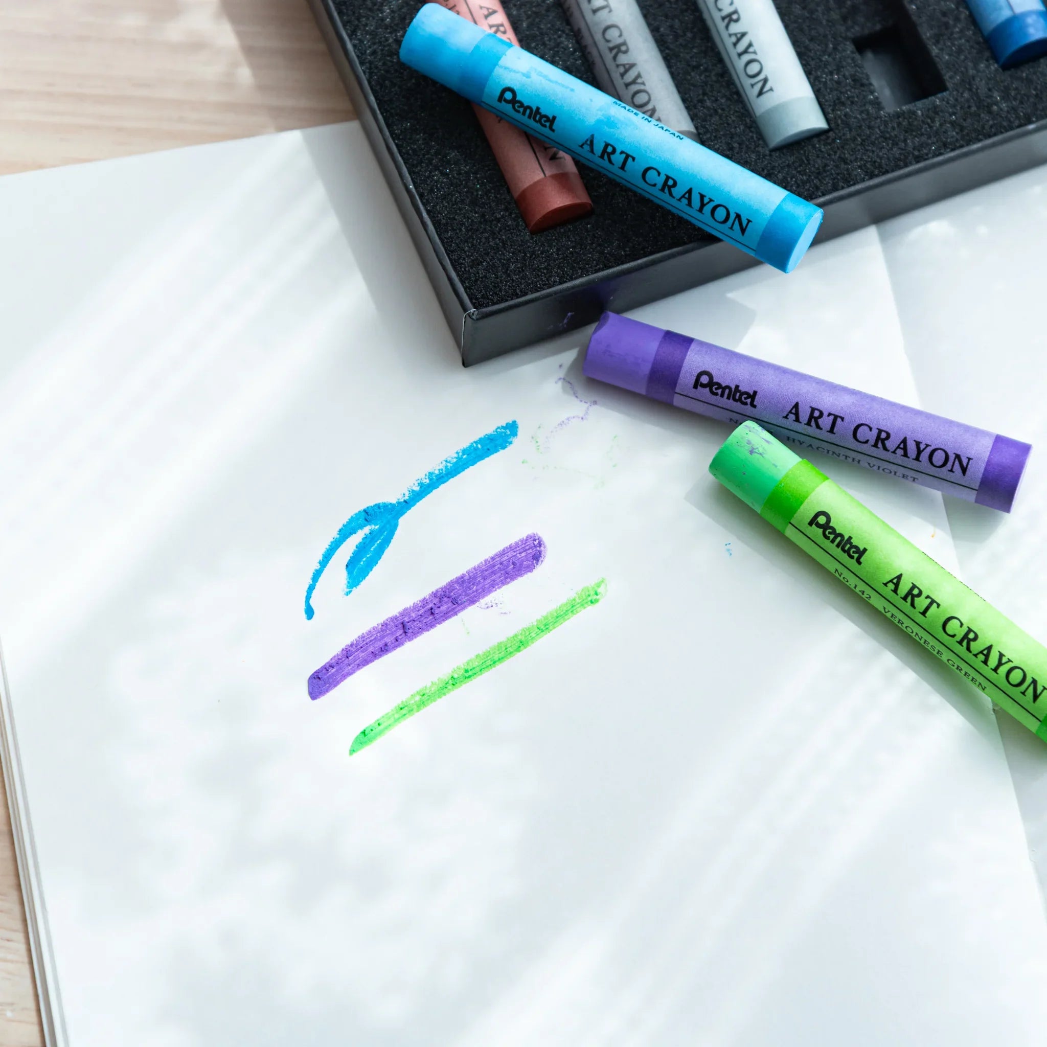 Three Pentel Art Crayons in sky blue, hyacinth violet, and veronese green on white paper with matching color stroke marks