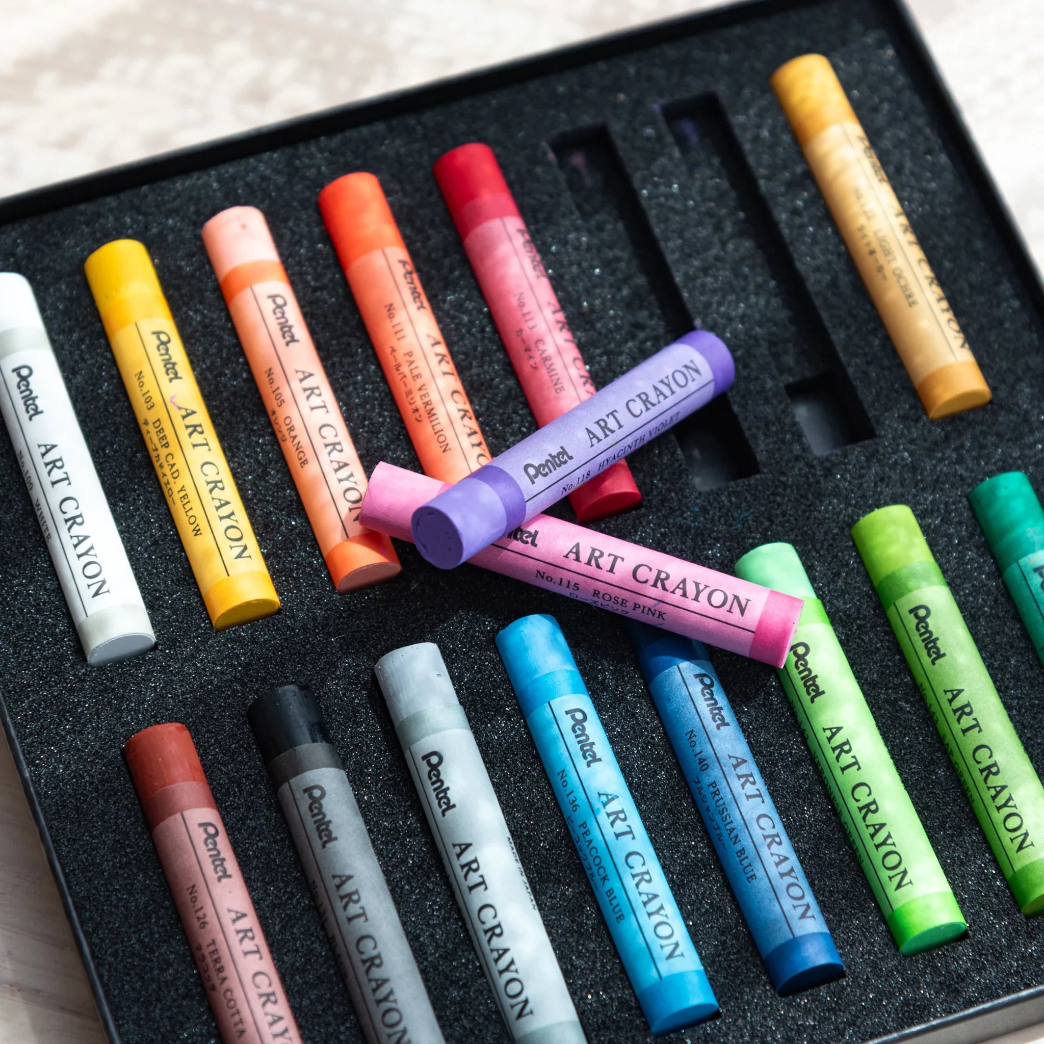Japanese crayon set by Pentel showing colorful art crayons in a foam tray with a pink crayon laid diagonally across other crayons