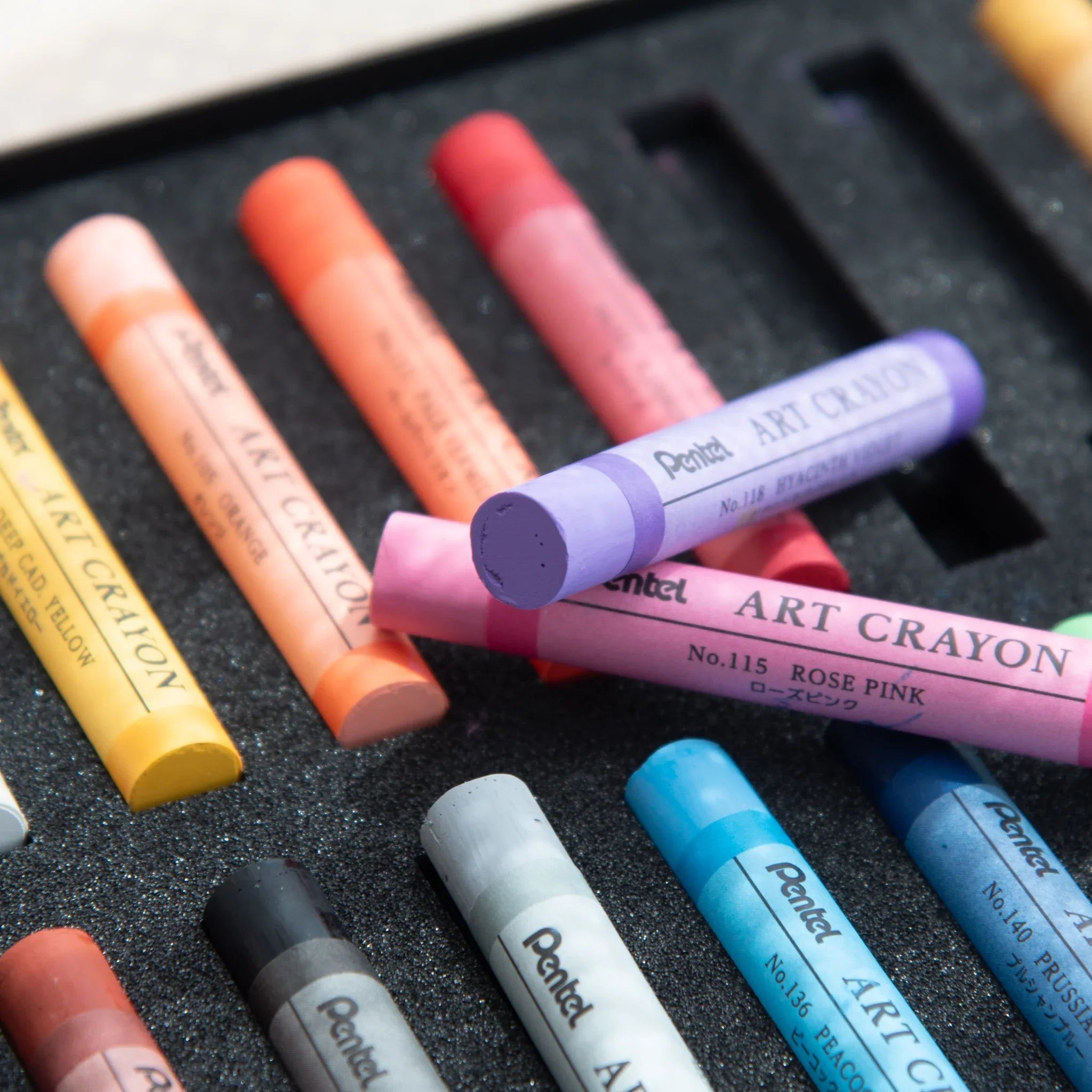 Pentel Art Crayons scattered on foam tray with Rose Pink No.115 and Hyacinth Violet No.118 overlapping in the foreground
