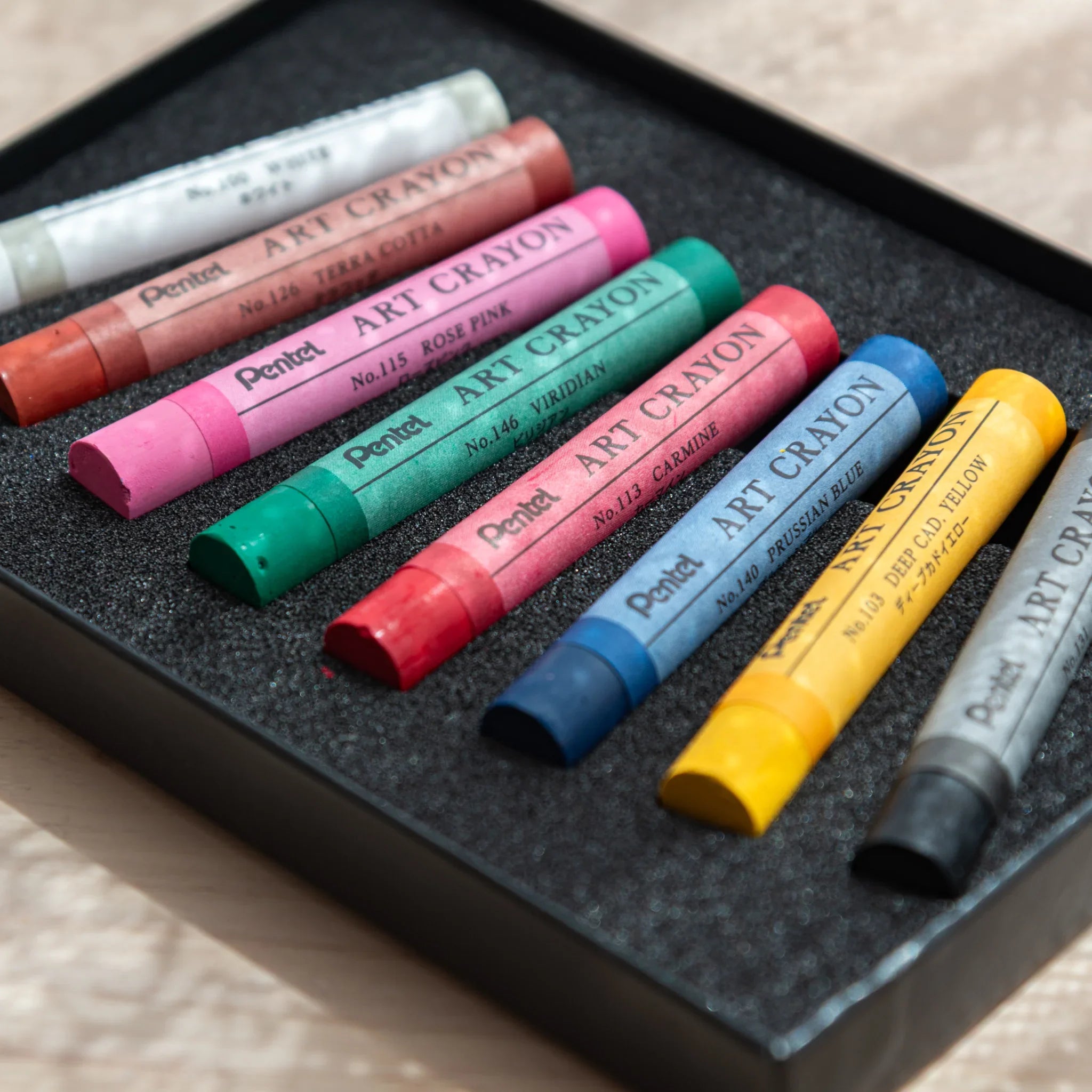Eight Pentel Art Crayon sticks spread in open black foam tray showing all labeled colors from black to white