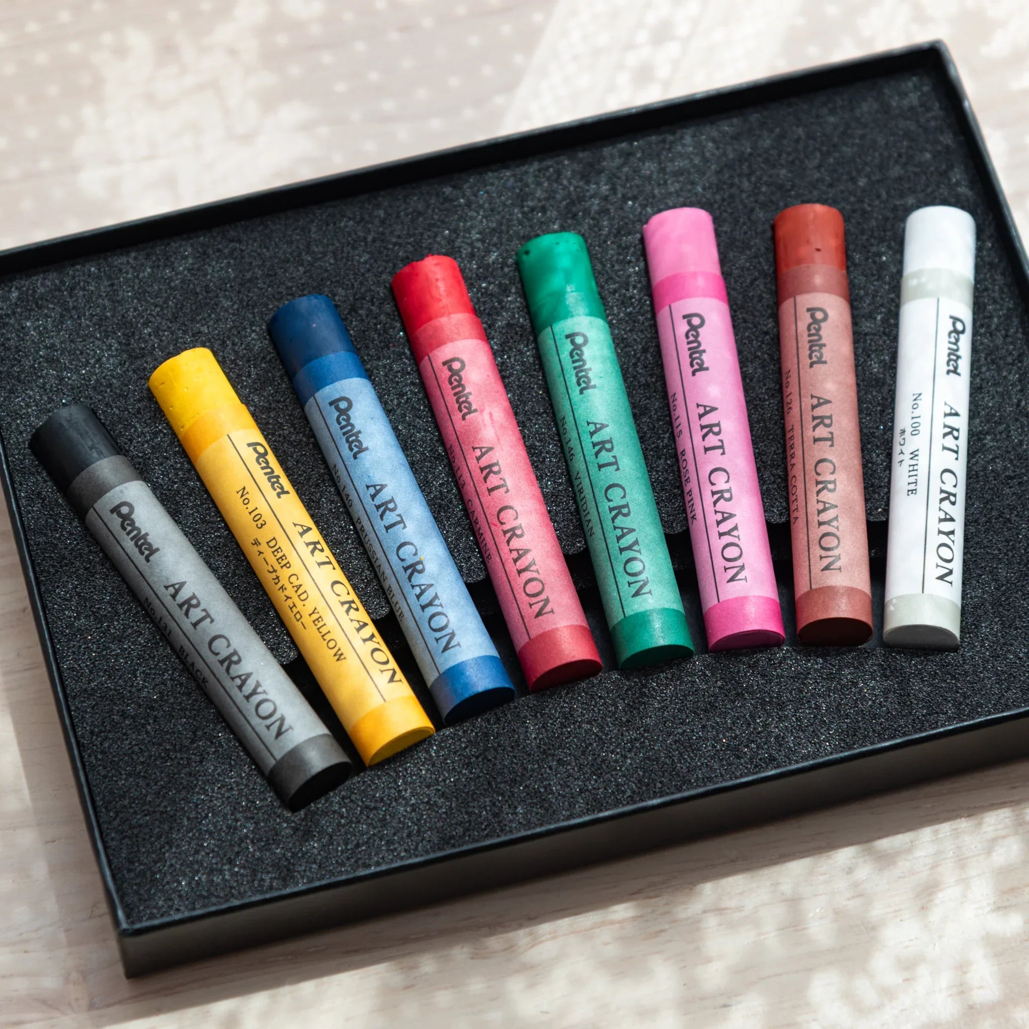 Eight Pentel Art Crayons fanned diagonally in open black foam box tray showing all colors from black to white