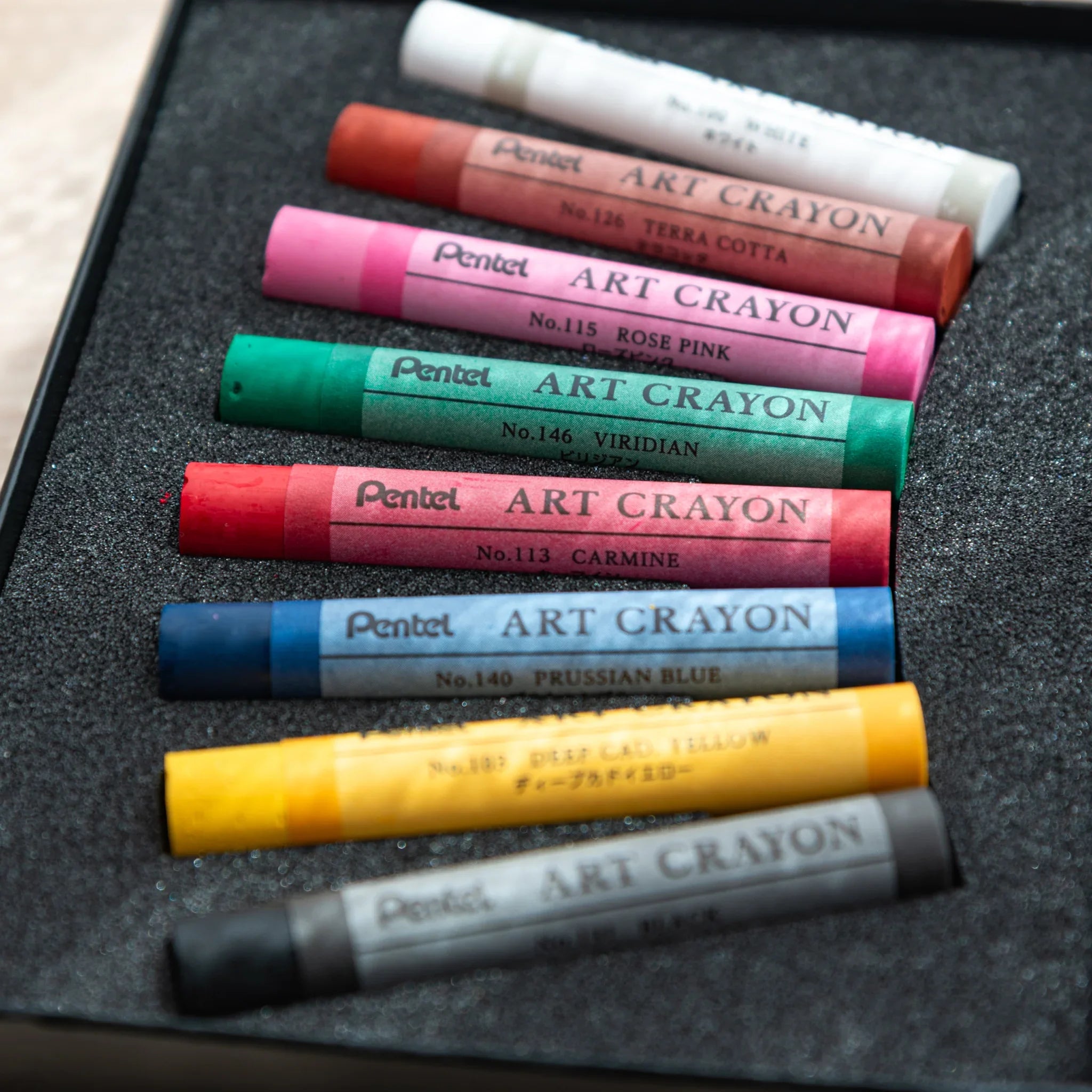 Eight Pentel Art Crayons showing label side in black foam tray with names including Prussian Blue, Viridian, and Carmine