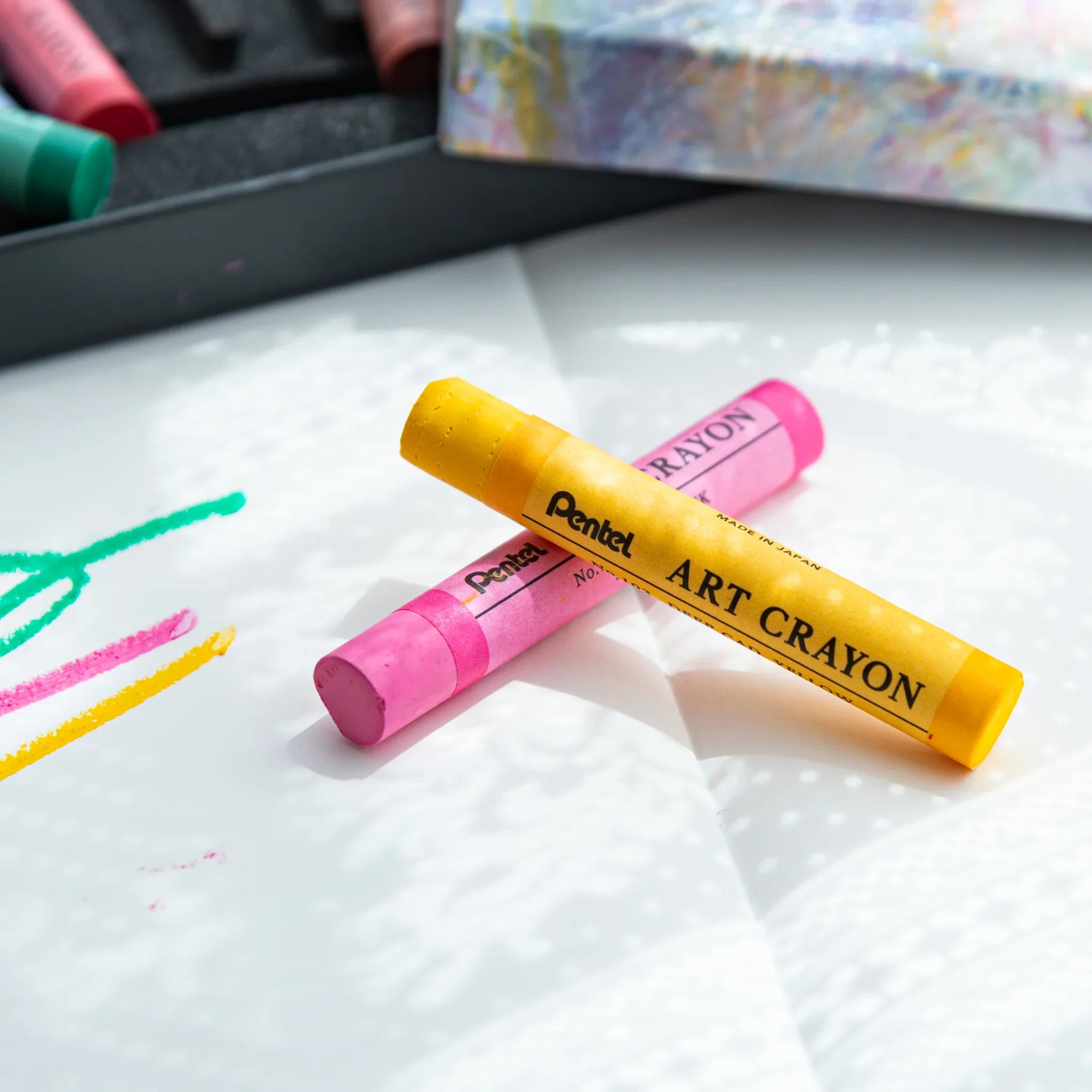Yellow and pink Pentel Art Crayons crossed on paper with green and yellow lines; Japanese crayon set by Pentel
