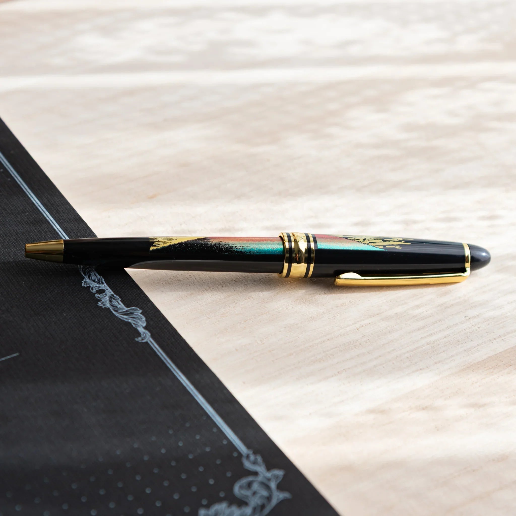 Japanese Ballpoint Pen with red Mt. Fuji and gold cloud design on a black notebook over a light wood desk.