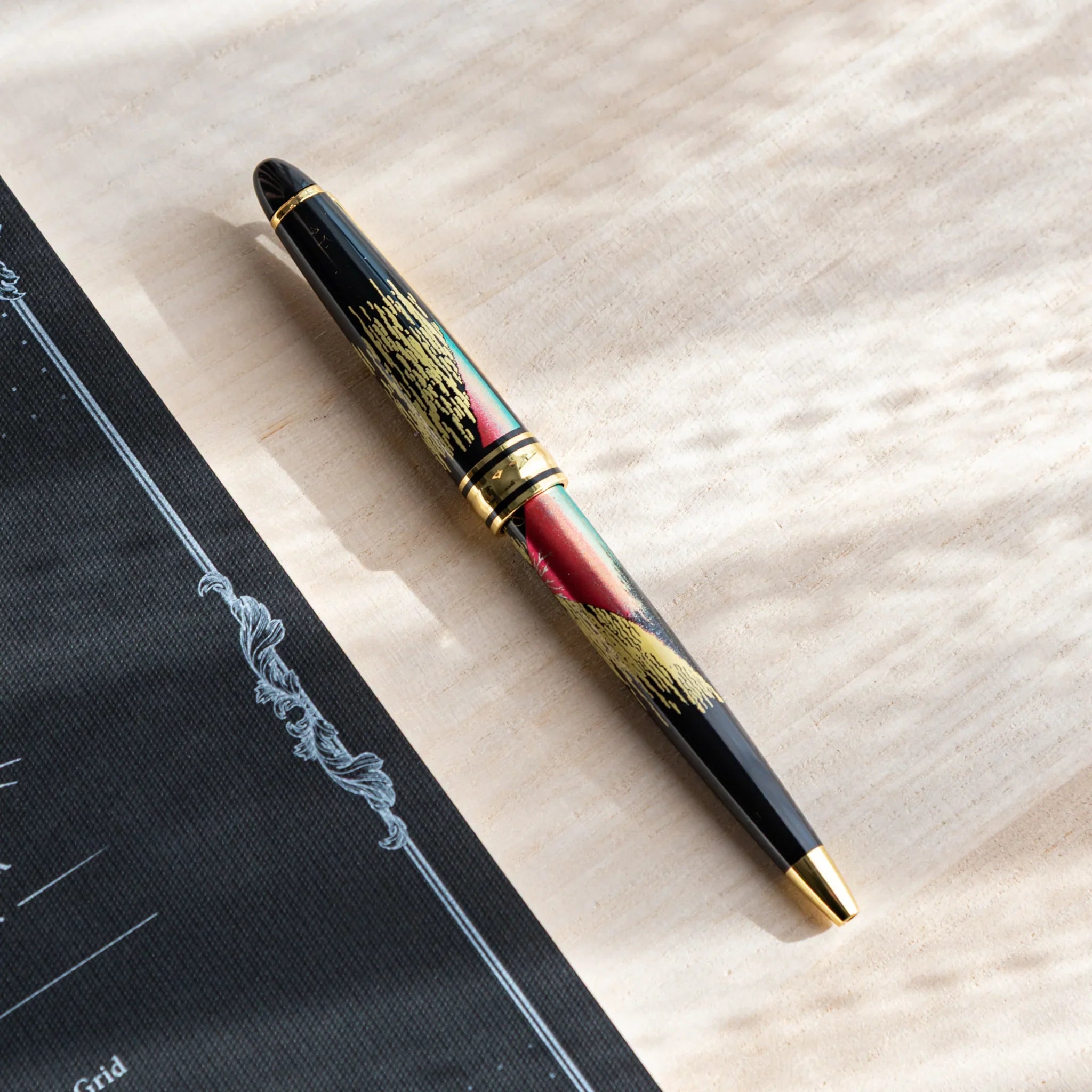 Japanese Ballpoint Pen with red Mt. Fuji and gold cloud design on a light wood desk beside a black notebook.