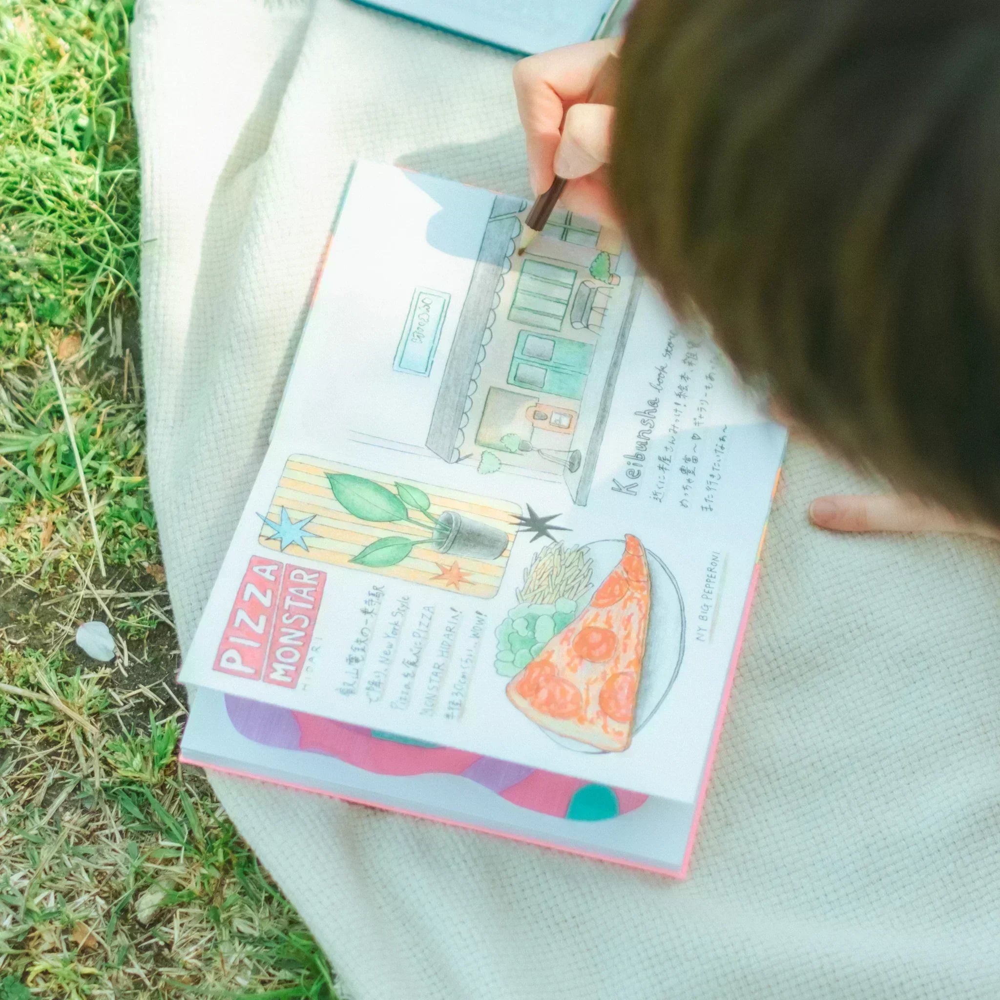 A woman sketching and coloring in a goshuincho notebook outdoors, drawing a pizza shop and food items on a sunny day.