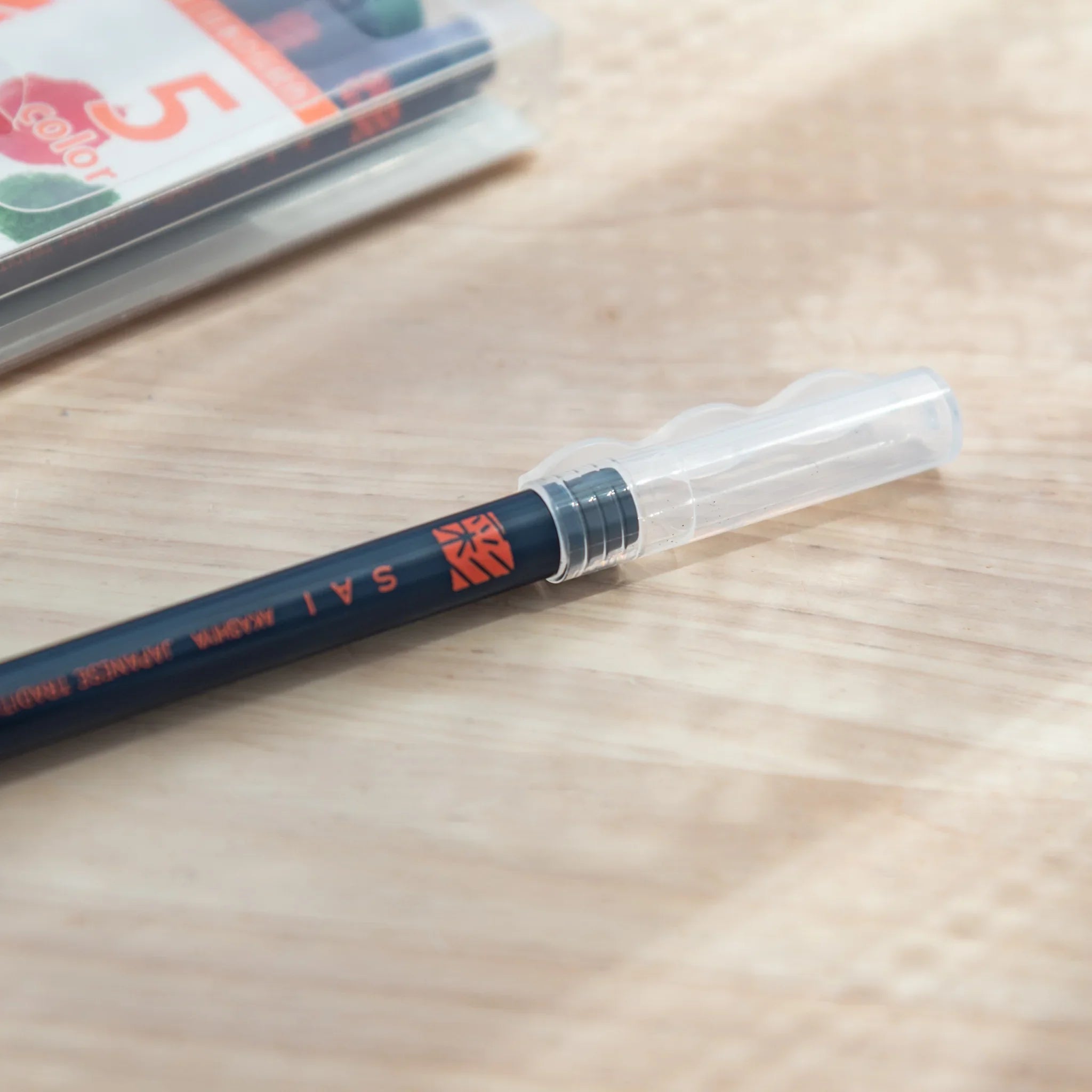 Akashiya SAI brush pen with clear cap removed, lying on wood surface showing dark navy barrel with orange SAI logo text