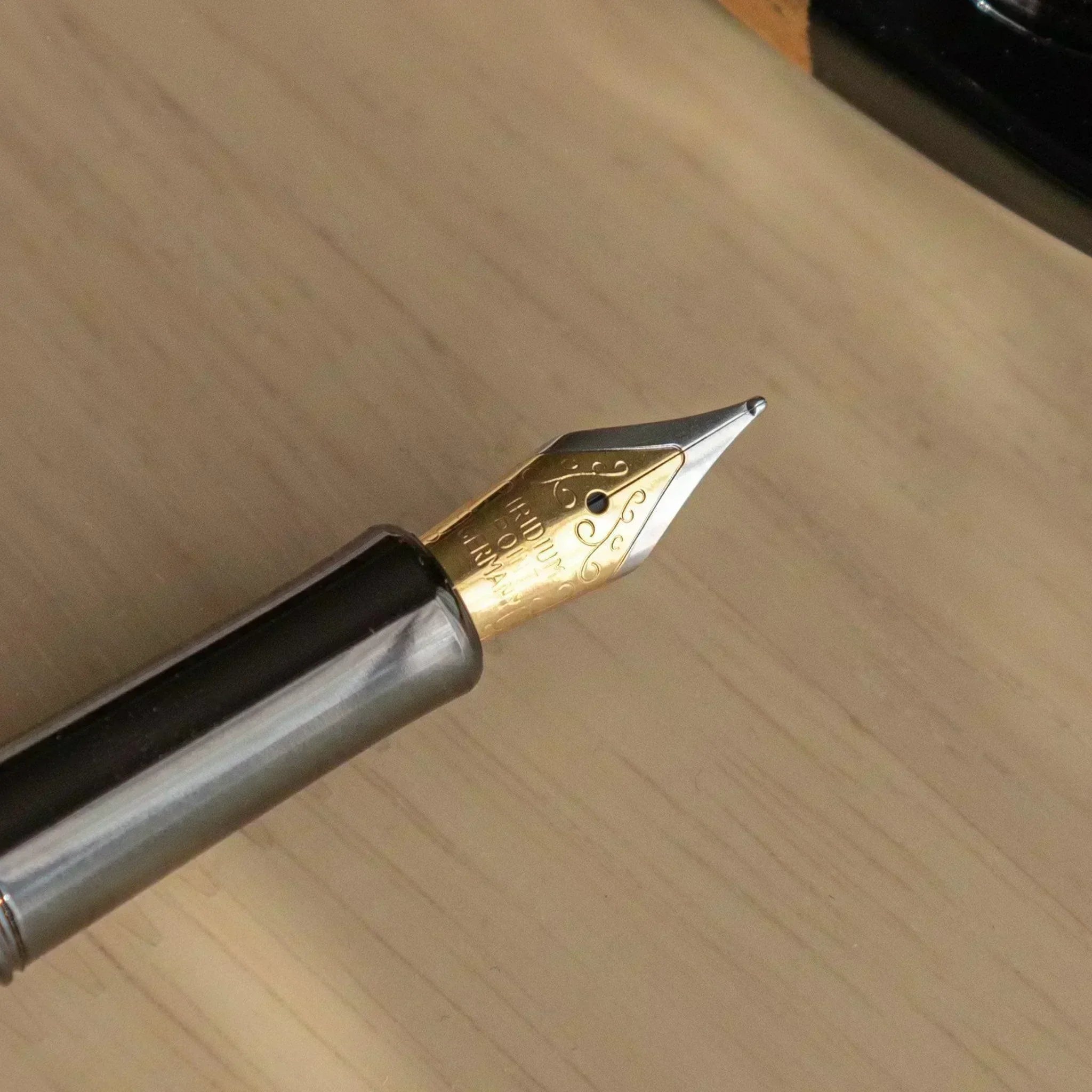 Close-up of a gold fountain pen nib with engraved details, resting on a light wooden desk, showcasing fine craftsmanship.
