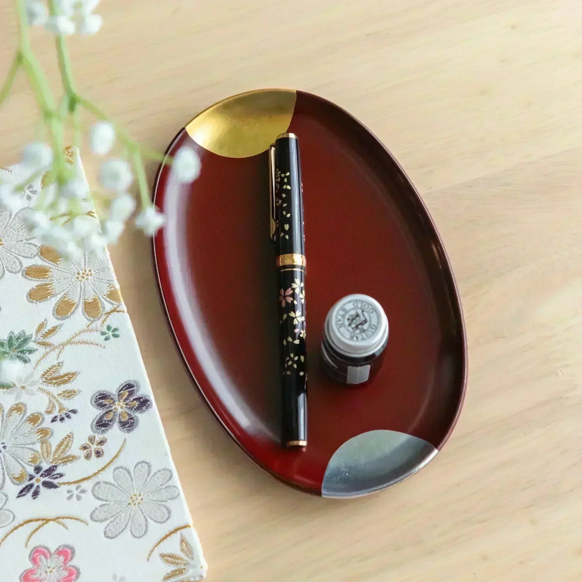 A Japanese stationery setup featuring a black sakura pen and ink bottle on a red lacquer tray with gold and silver accents.
