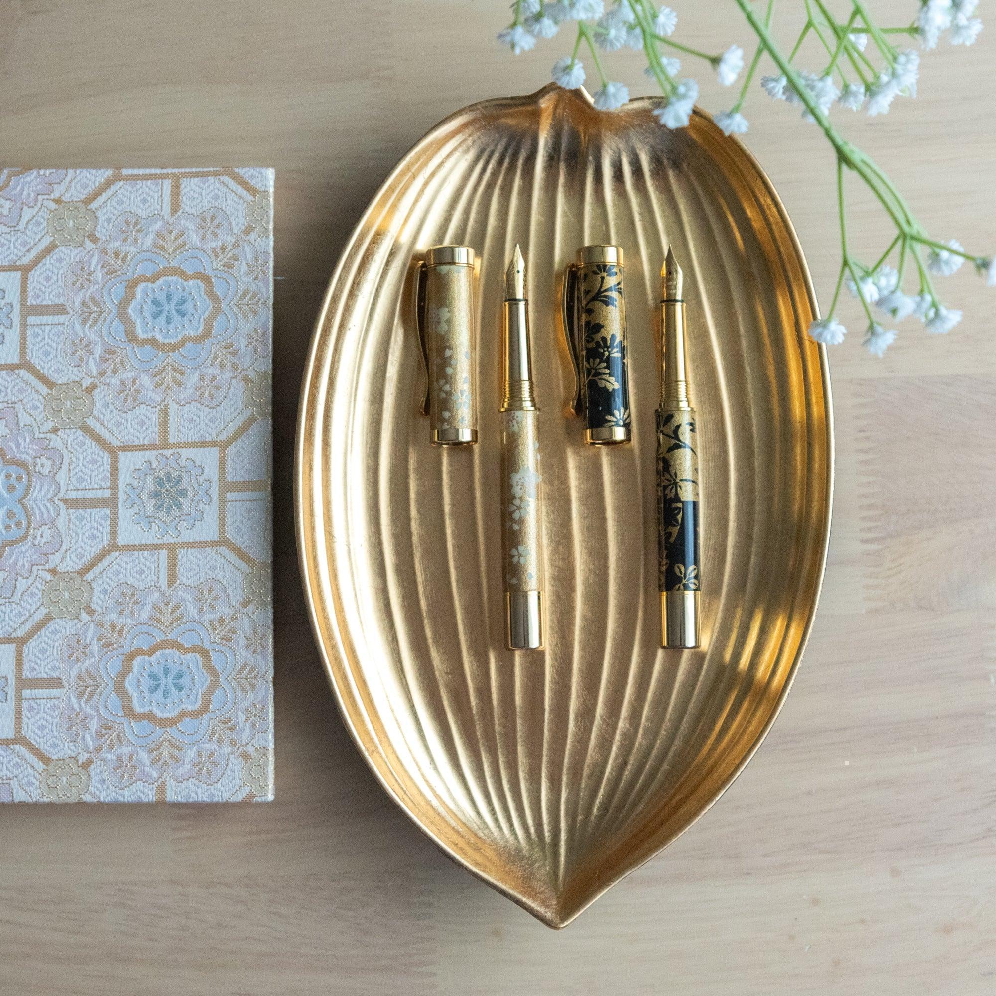 Two elegant Japanese stationery fountain pens with floral designs displayed on a gold leaf-shaped tray.
