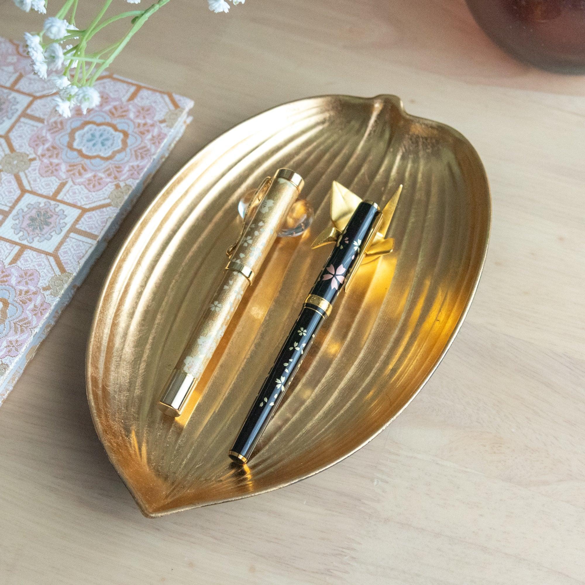 A black sakura-patterned fountain pen and a gold pen on a golden leaf tray, paired with a decorative origami.