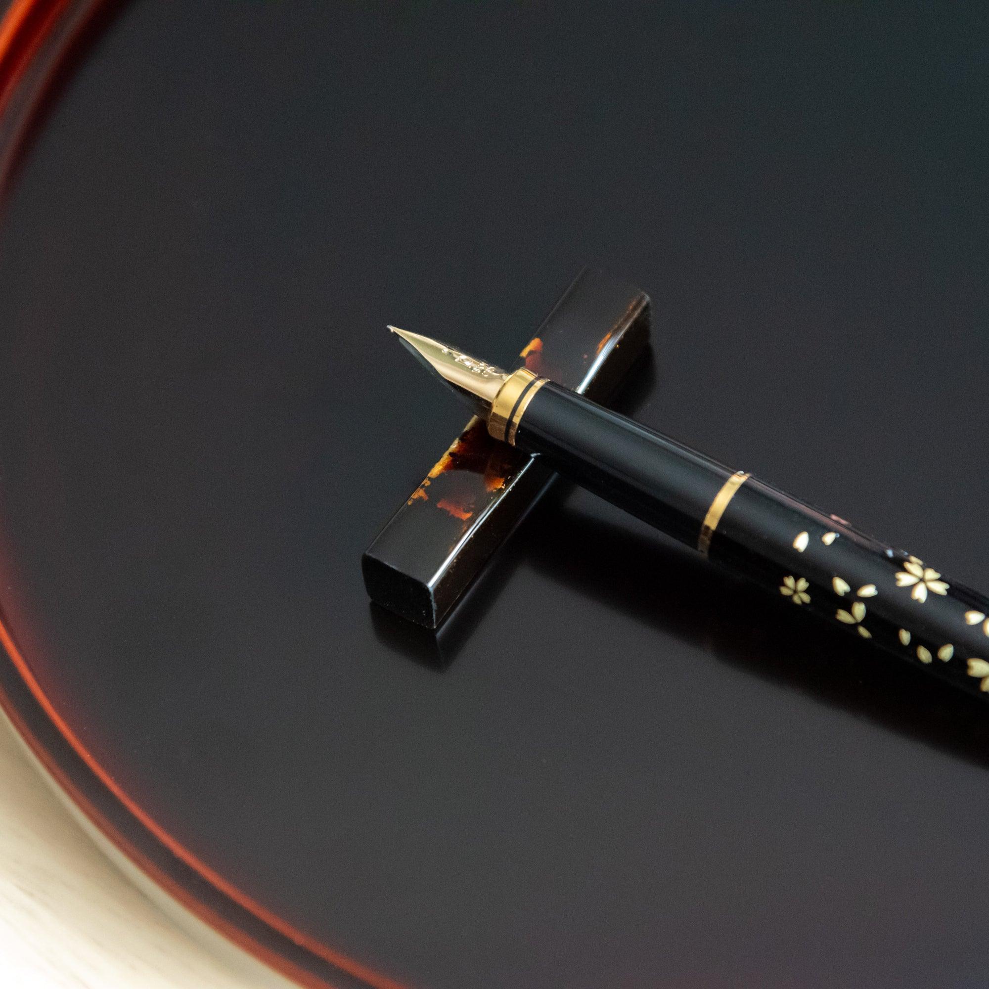 A Japanese stationery fountain pen with a sakura design rests on a black and red marbled pen rest on a glossy black tray.