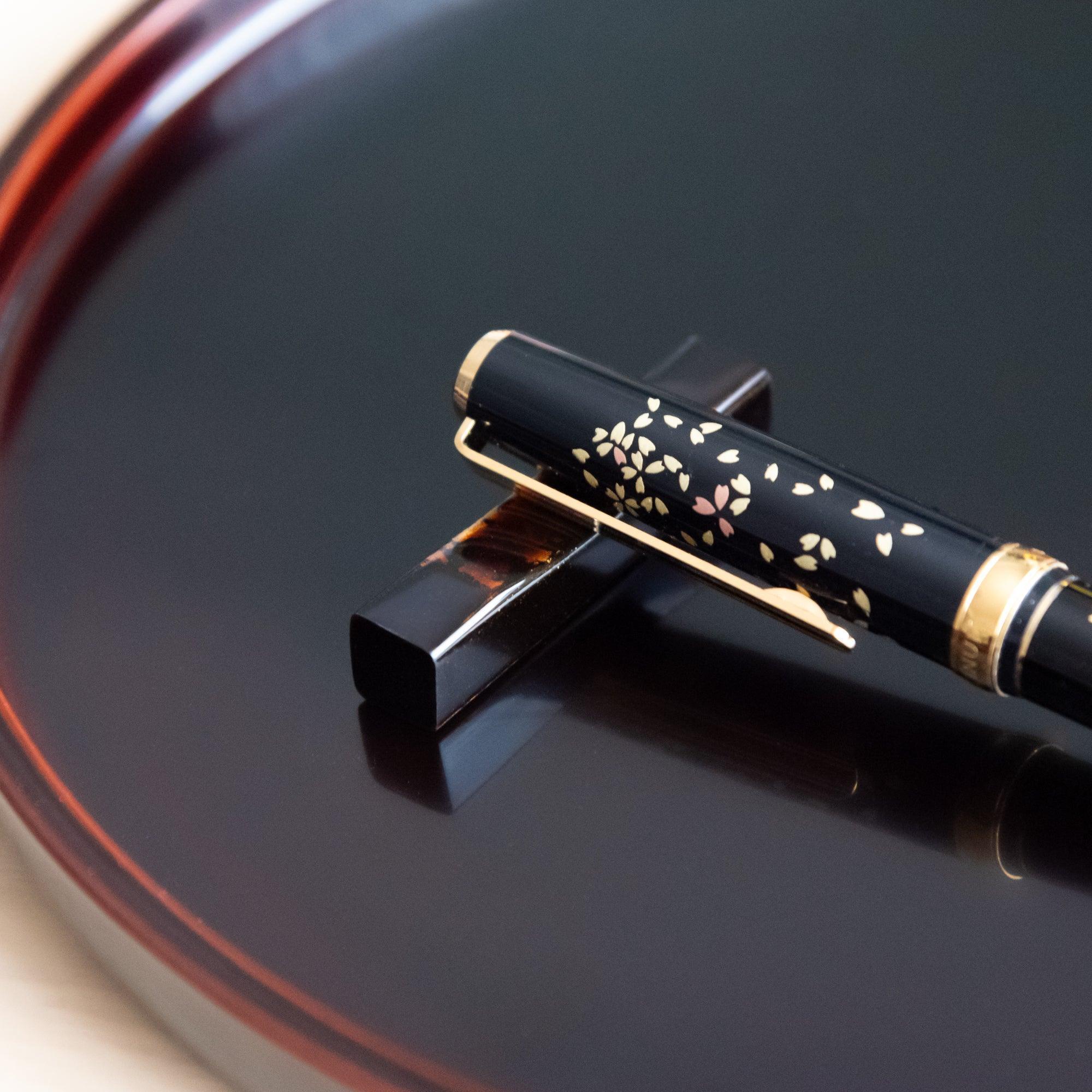 A close-up of a Japanese stationery pen on a black and red marbled pen rest, emphasizing its floral sakura pattern.