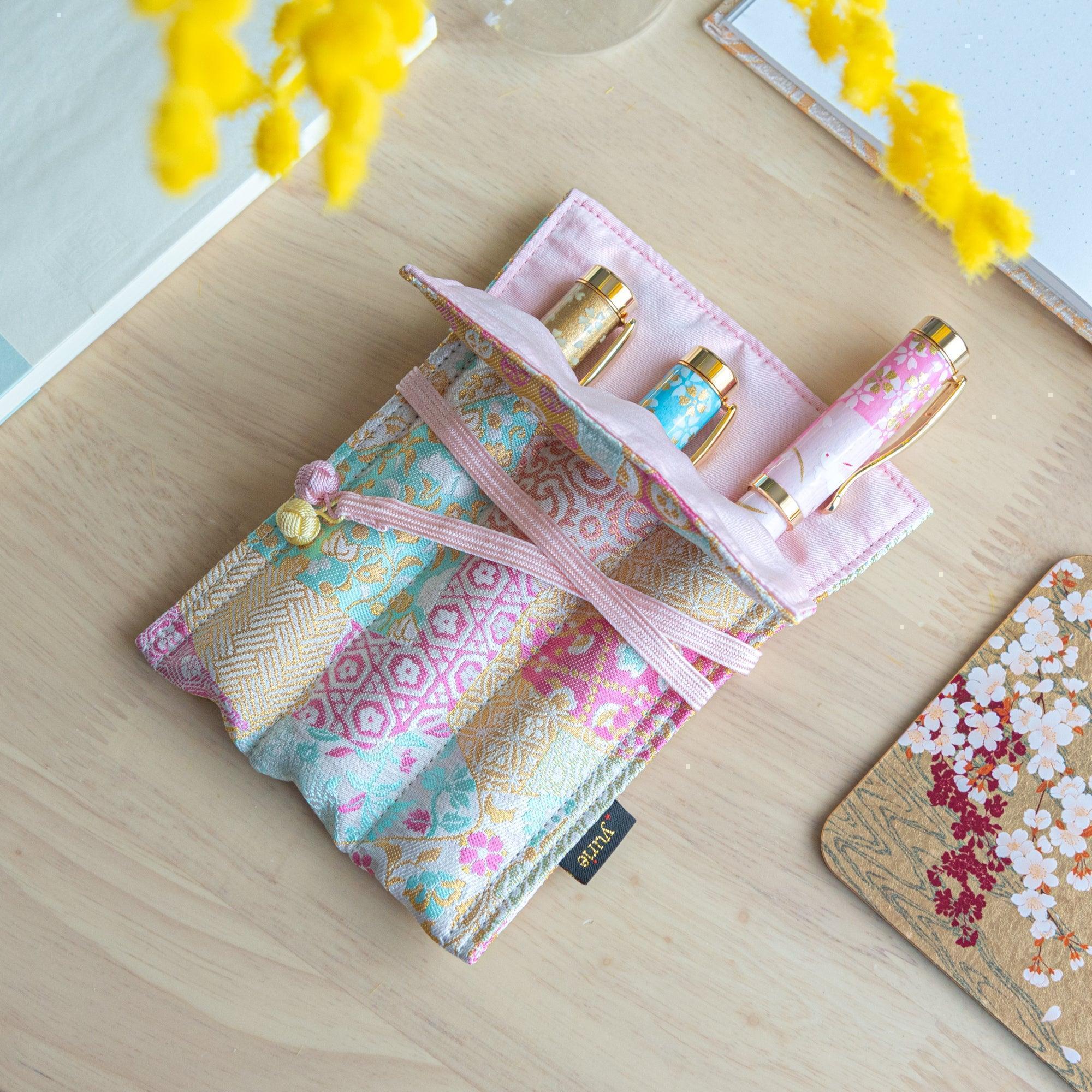 A pastel-colored Japanese fountain pen case of Nishijin silk, with a pink interior holding three decorative fountain pens on a wooden desk.