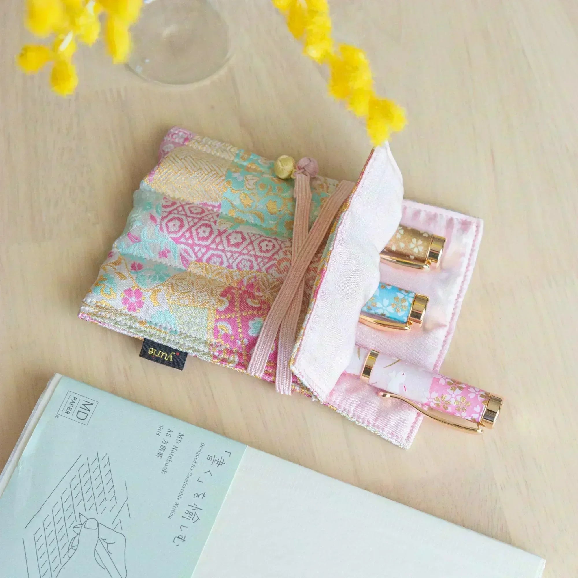 A pastel-colored Japanese fountain pen case, partially open to reveal three ornate fountain pens, surrounded by stationery and mimosa flowers.