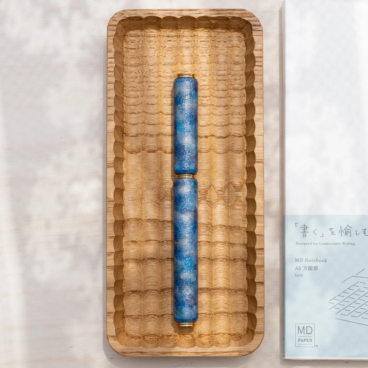 Blue marbled urushi lacquered rollerball pen in zelkova wood, shown in a carved wooden tray with a Japanese notebook