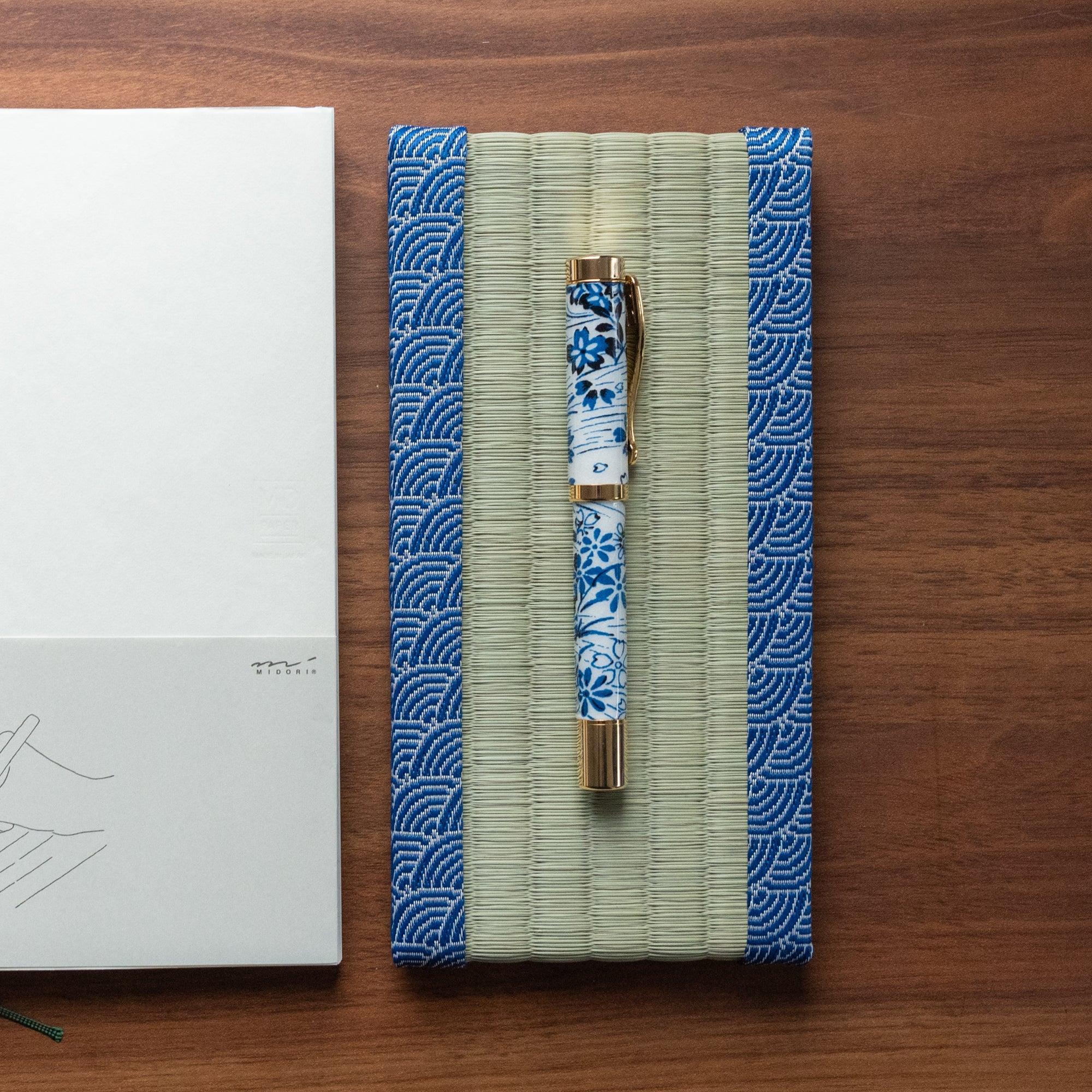 Blue and white floral fountain pen on a tatami pen tray with traditional Japanese patterns, alongside a minimalist notebook, showcasing elegant Japanese stationery.