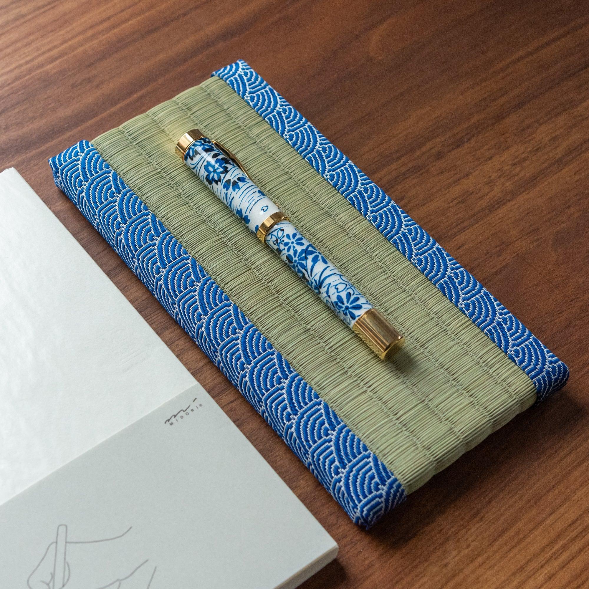 Angled view of a floral fountain pen resting on a tatami pen tray bordered with blue wave patterns, paired with a notebook, representing Japanese stationery.