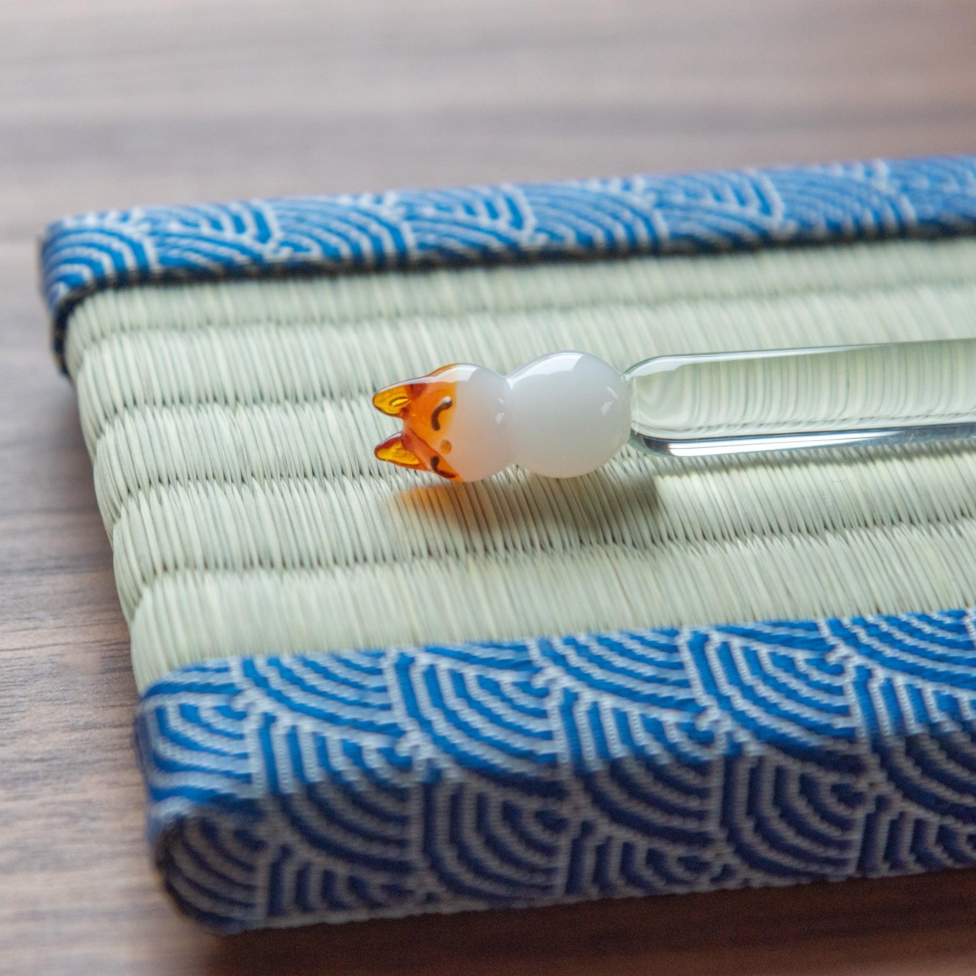 Close-up of a glass pen with a fox motif resting on a tatami pen tray, emphasizing the detailed artistry and cultural essence of Japanese stationery.
