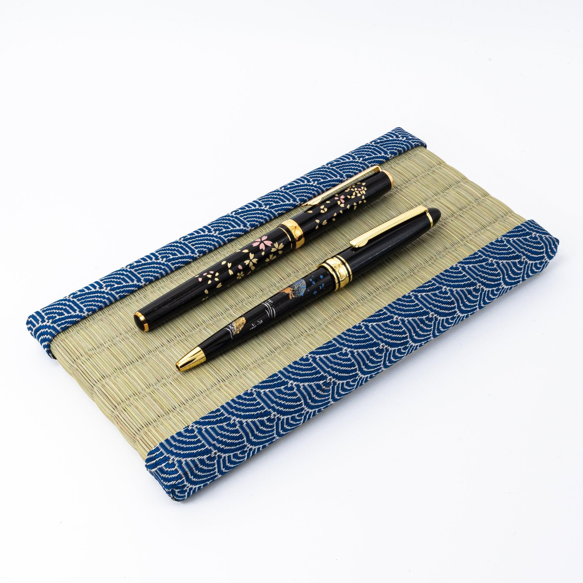 Two black lacquer pens with gold floral and wave motifs resting on a tatami pen tray with blue wave borders, emphasizing refined Japanese stationery.