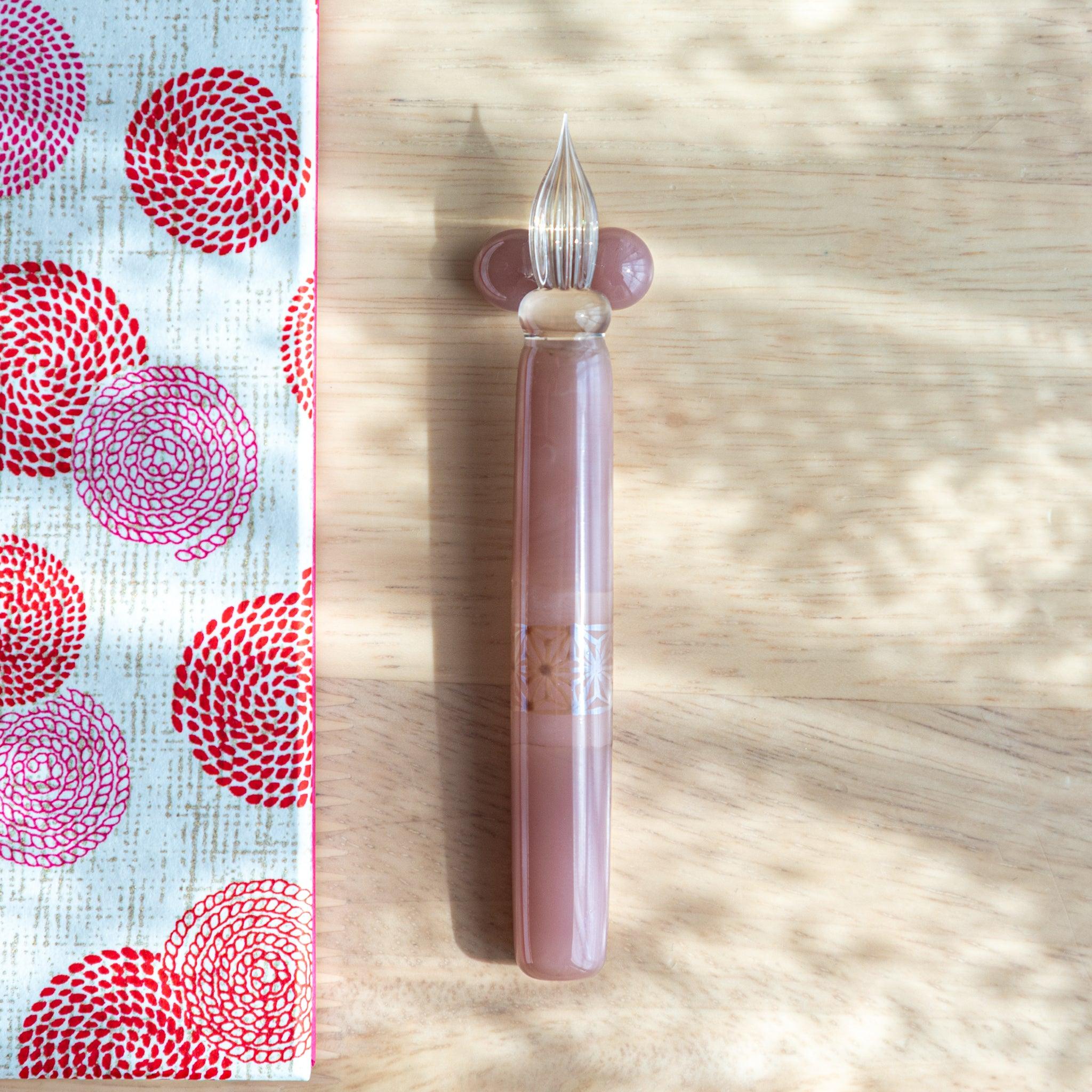 Top-down view of a Japanese pink glass dip pen with a hemp leaf motif and transparent nib, placed next to a red swirl notebook.