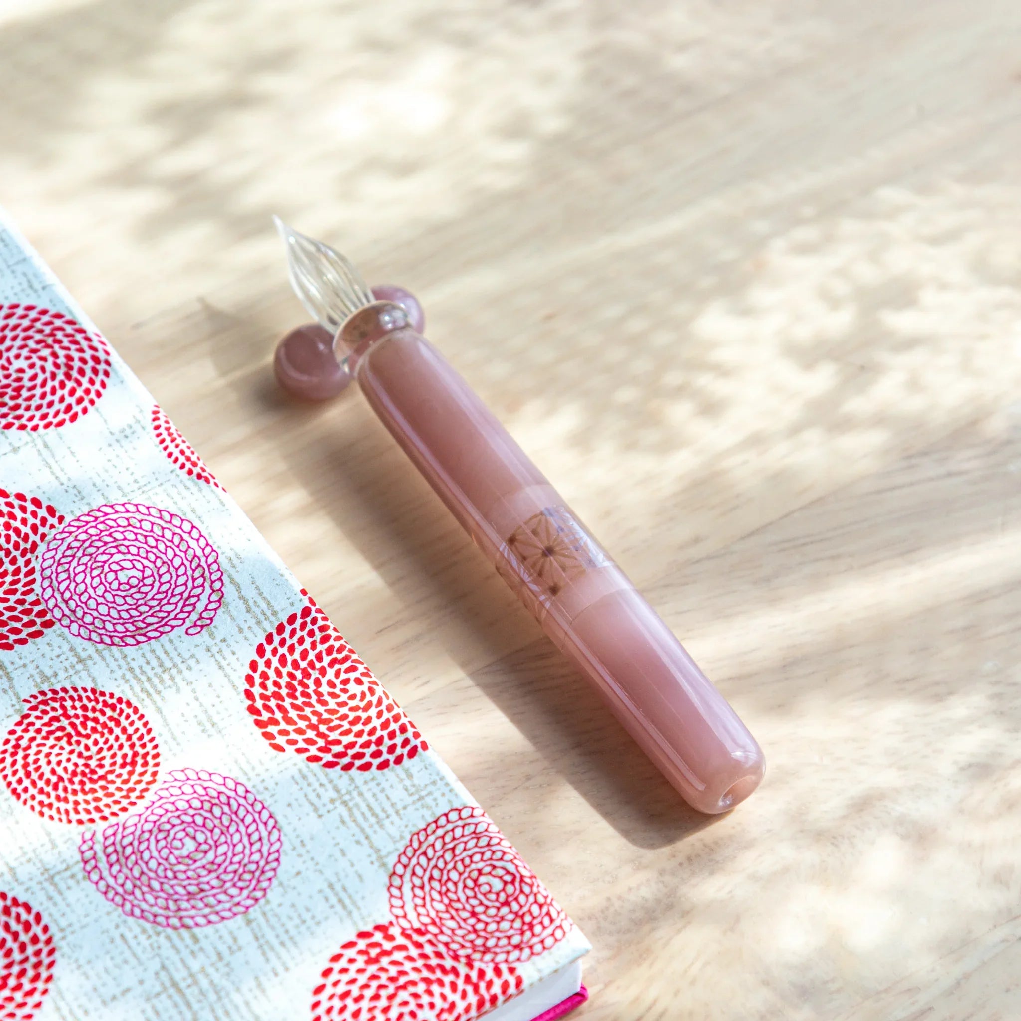Blush pink glass dip pen with hemp leaves design shown on pink patterned notepad with wooden pen rests