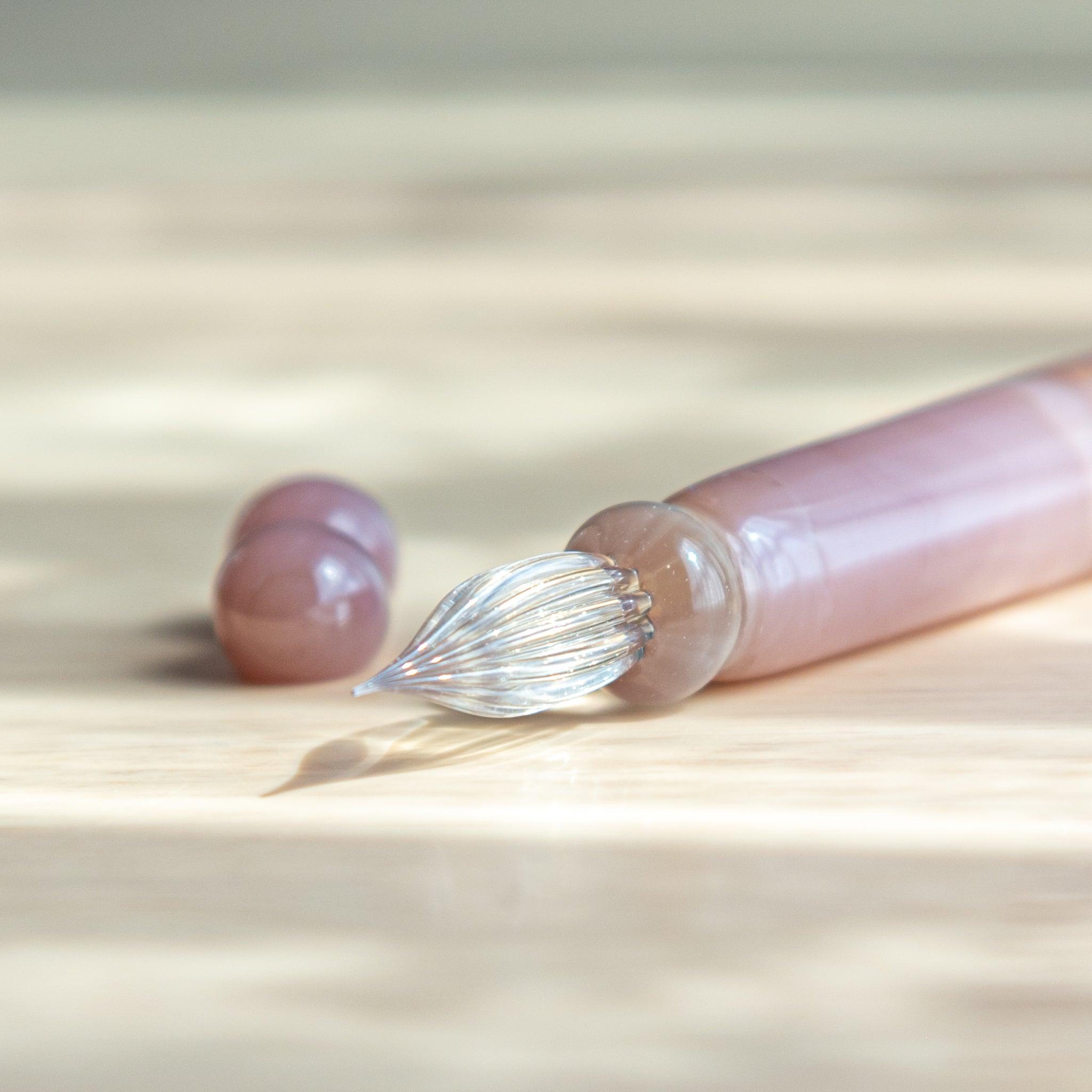 Close-up of a pink glass dip pen from Japan, showing its twisted clear nib and glossy body with hemp leaf engraving.