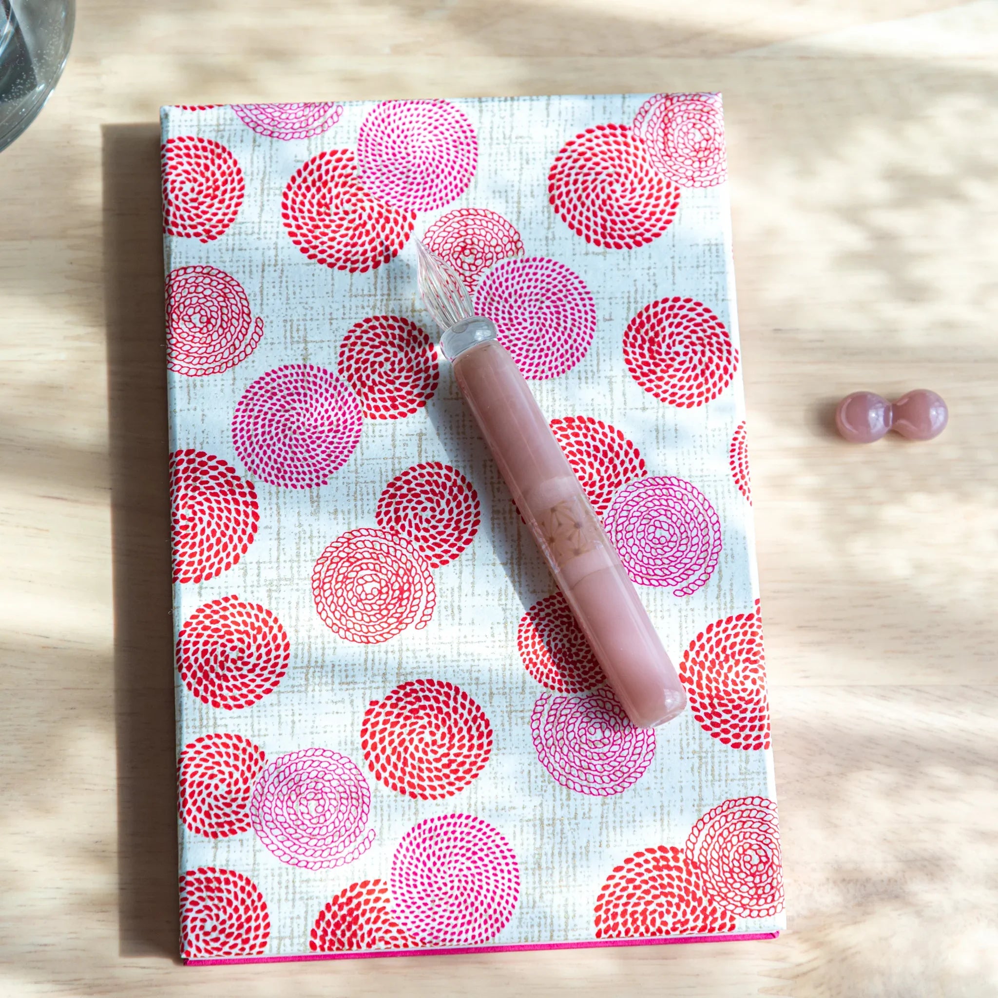 Japanese glass dip pen in blush pink with spiral nib placed on white notebook with circular hemp leaf print in red and pink