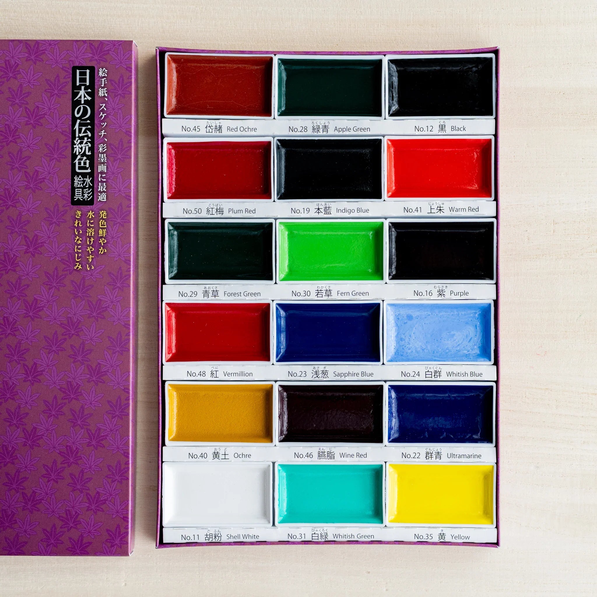 Open Gansai 18-color box from above showing eighteen dry watercolor pans in six rows of three, with bilingual labels