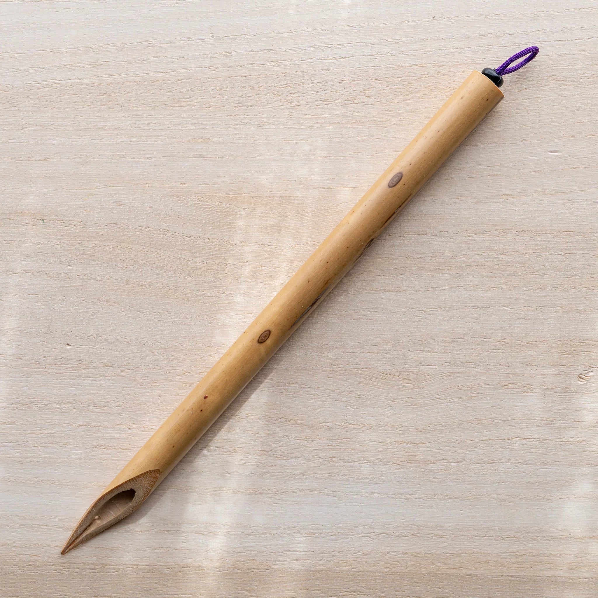Boku-Undo natural bamboo dip pen lying diagonally on wooden surface, pointed tip at bottom, purple hanging loop at top