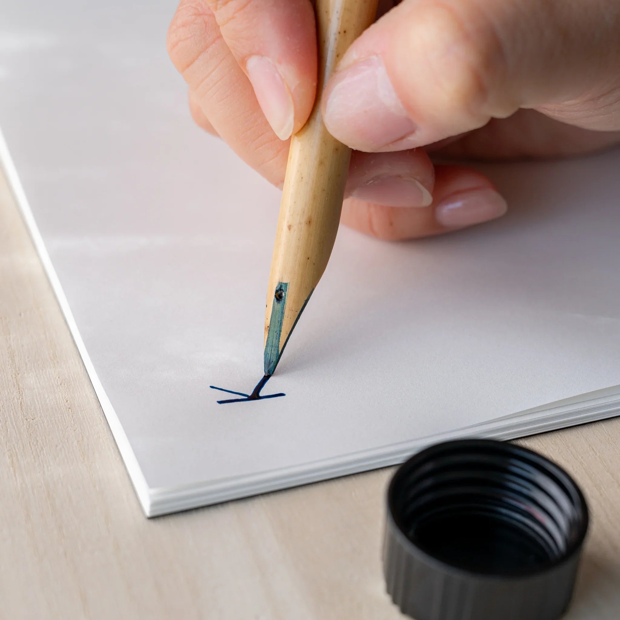Hand holding Boku-Undo bamboo dip pen writing dark blue ink strokes on white paper, black ink bottle cap in foreground