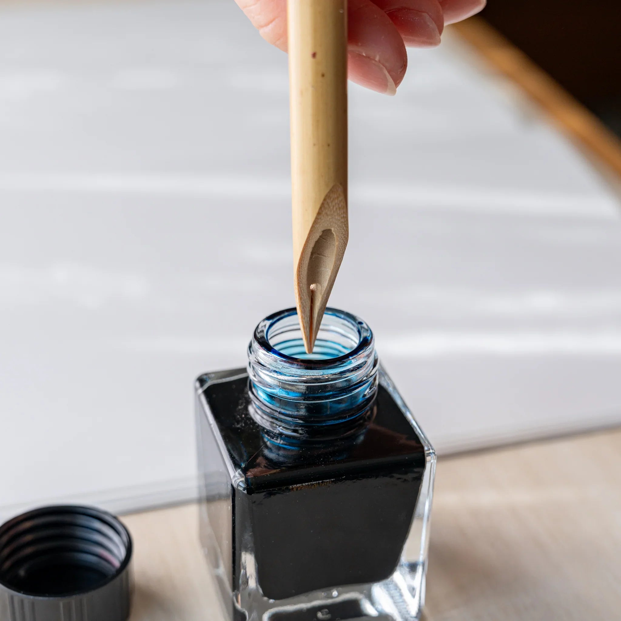 Hand dipping Boku-Undo bamboo dip pen into open ink bottle, showing the bamboo nib tip above dark ink, cap at lower left