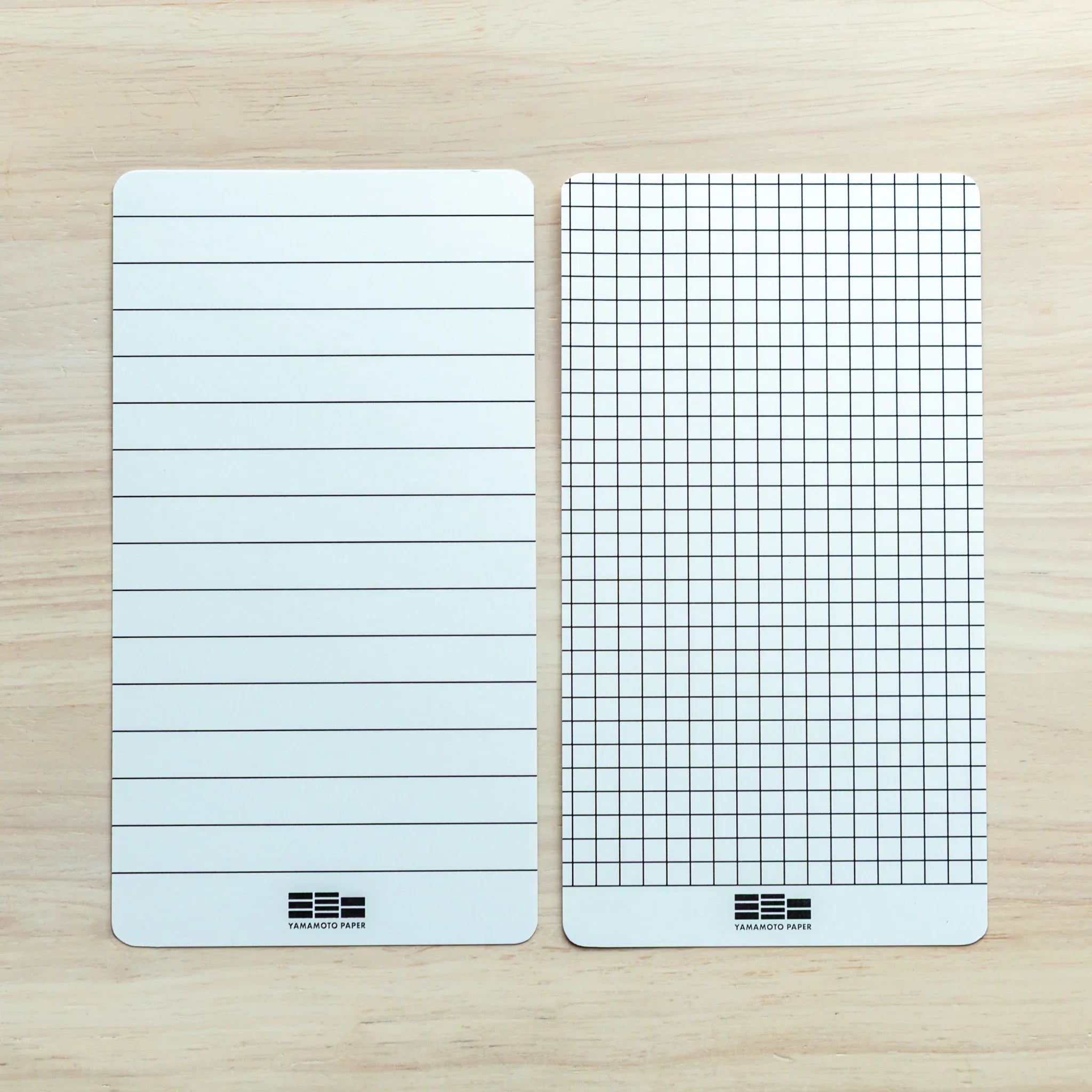 Two Yamamoto Paper pads on light wood: left with ruled lines, right with grid, Yamamoto Paper logos at bottom
