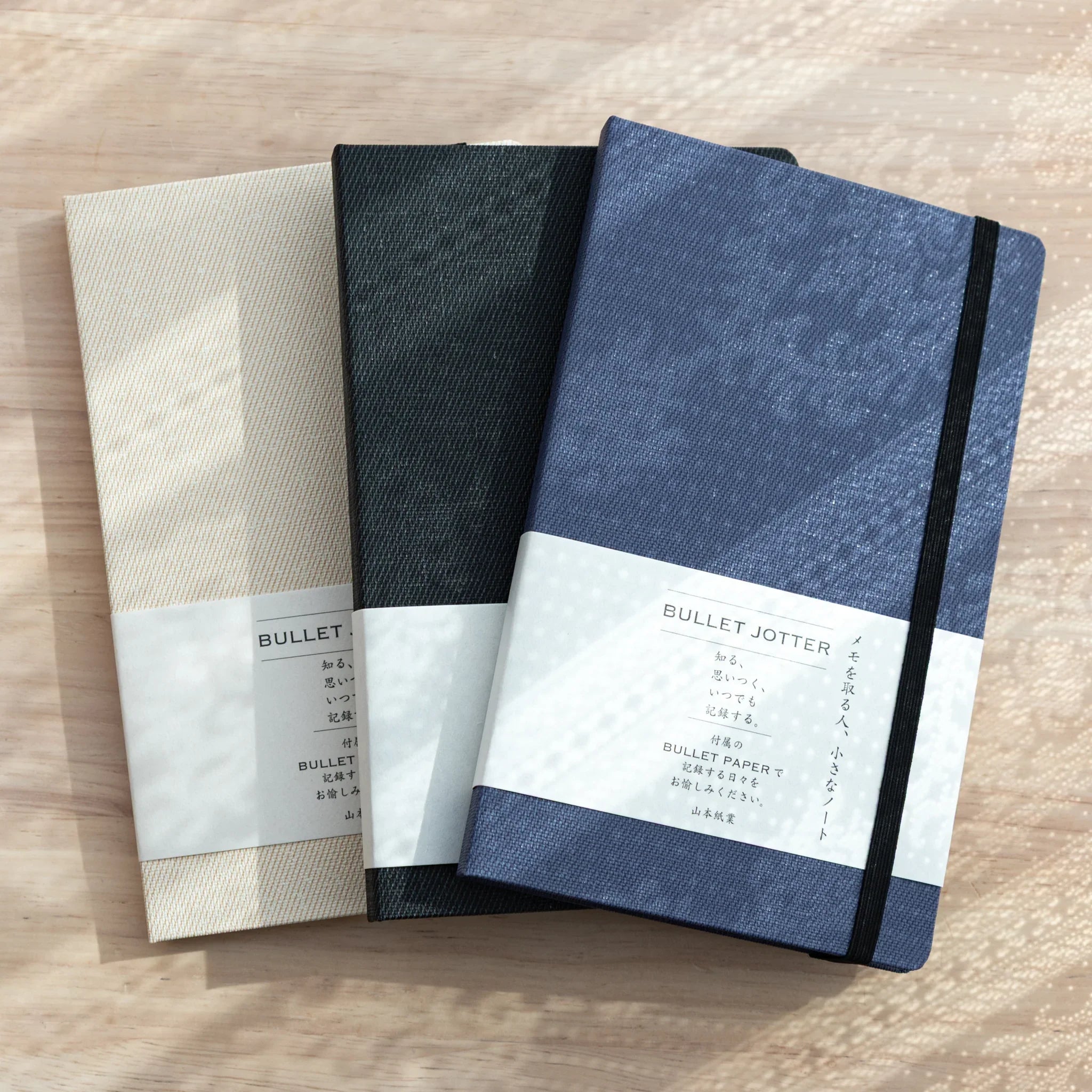 Three Bullet Jotter gabardine covers fanned out on wood: ivory white, dark gray, and royal blue, each with white belly band