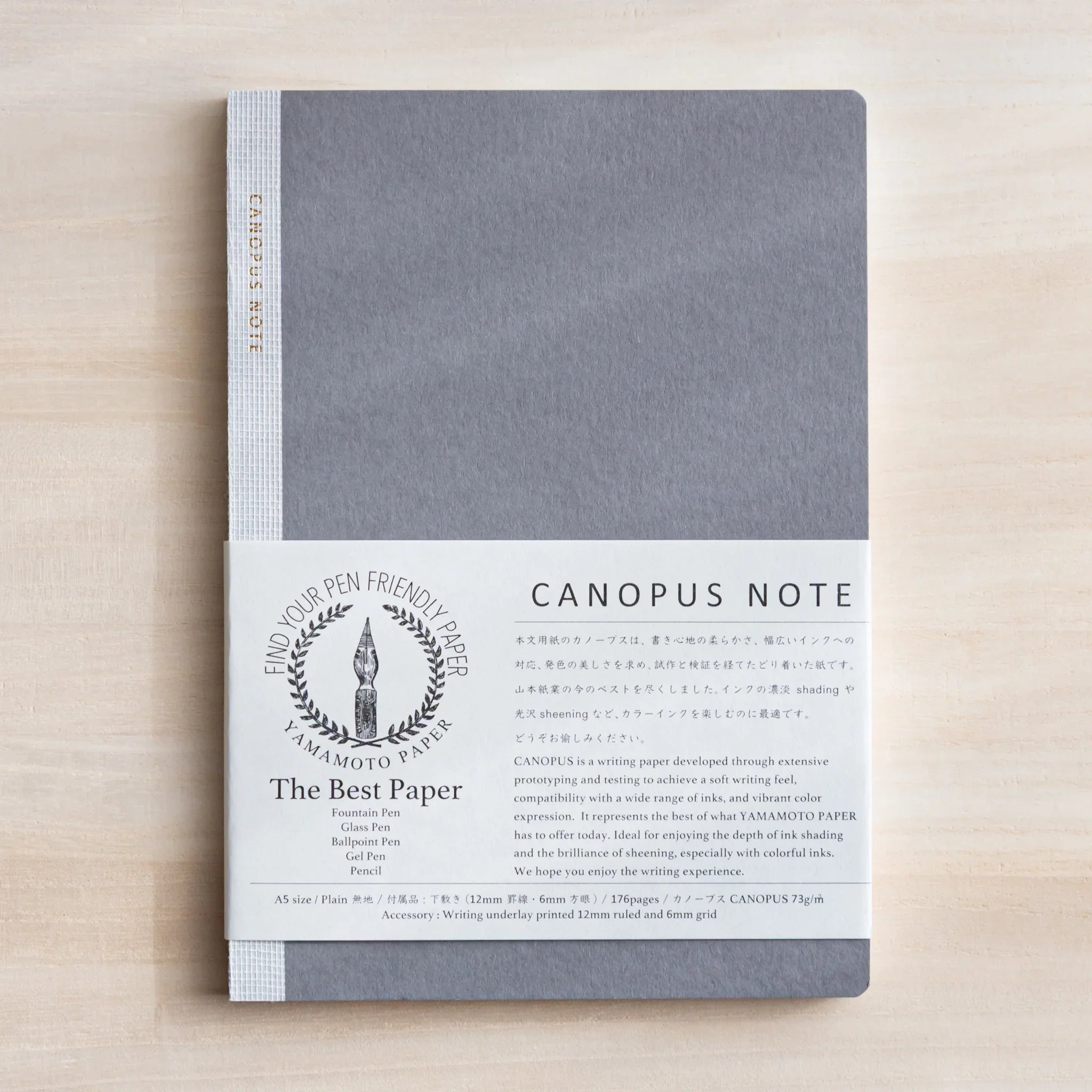 Closed grey Japanese Notebook with fabric cover wrapped in white band reading CANOPUS NOTE on light wooden desk.