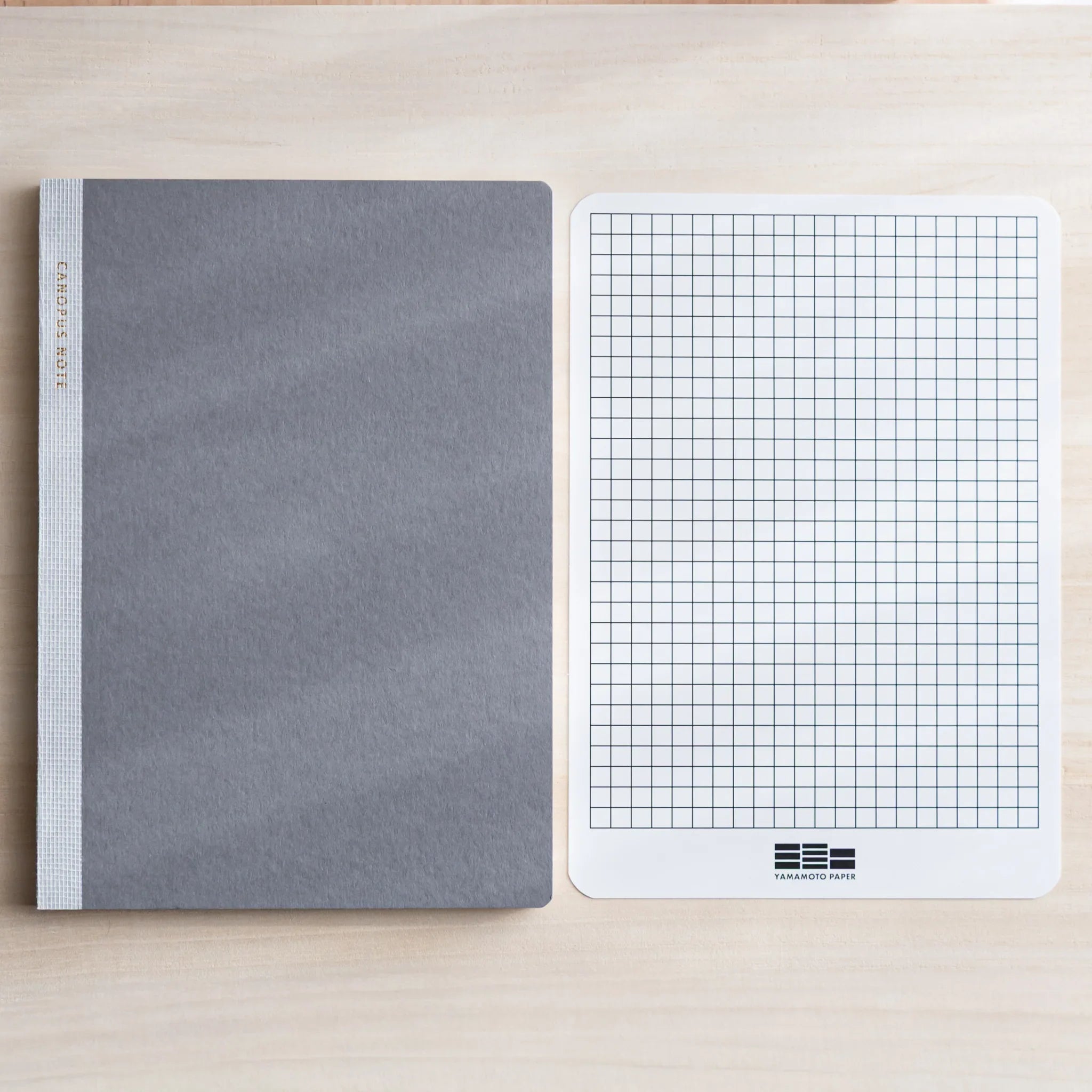 Closed grey Japanese Notebook beside white grid template sheet on light wooden desk.